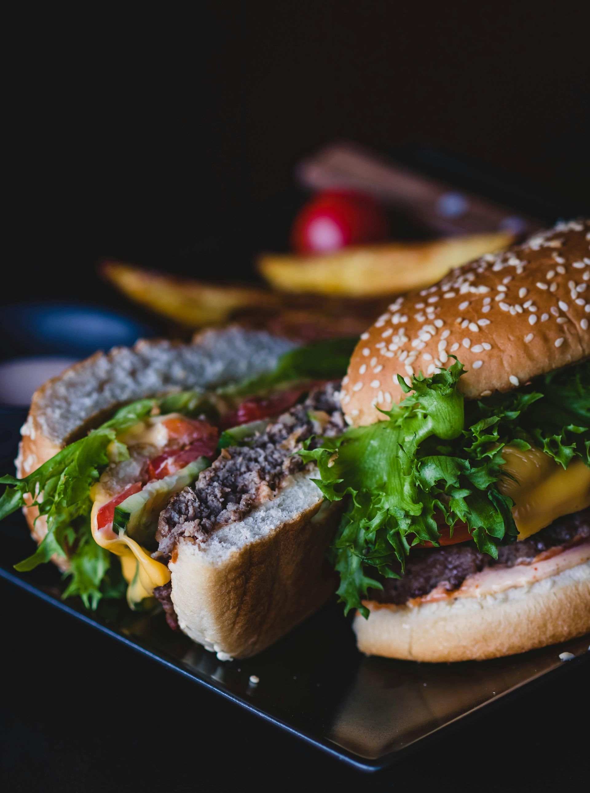 Hamburger cut in half, lettuce, tomato, cheese, sesame seed bun on a black plate, with potato wedges.