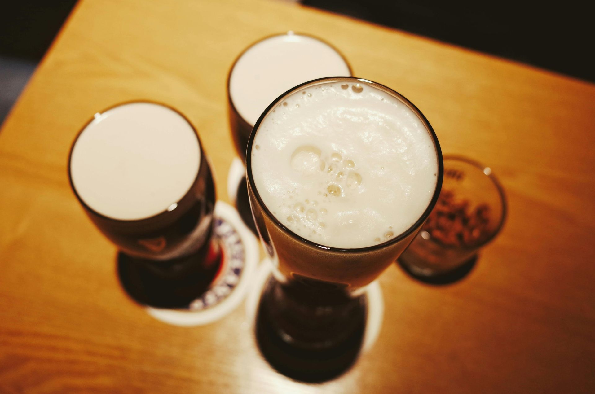 Four glasses of dark beer with foamy heads, on a wooden table, next to a small glass with nuts.