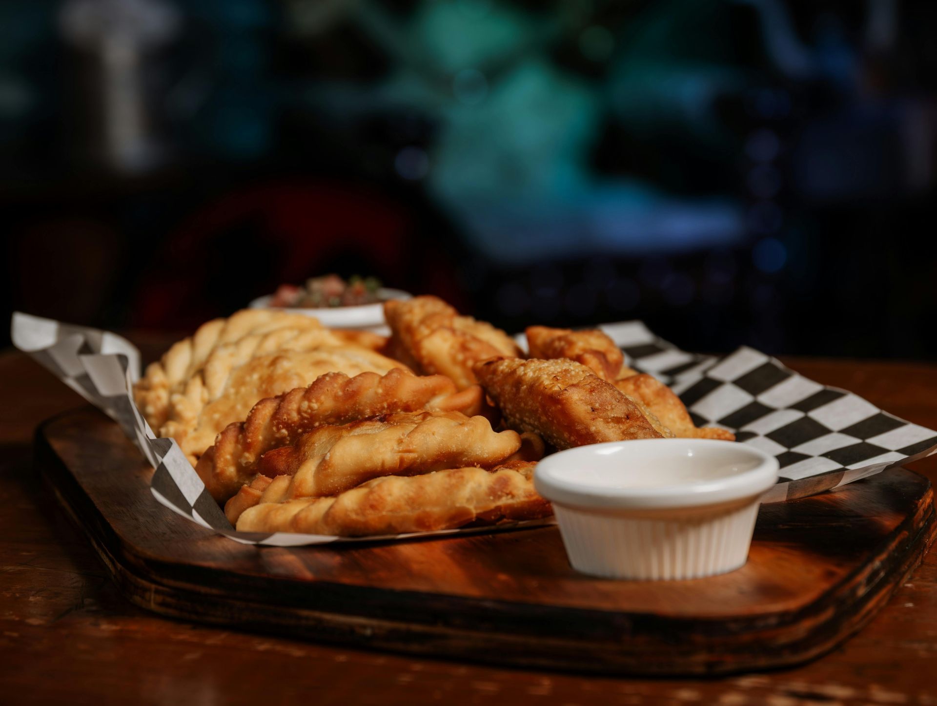 Fried empanadas and other fried items on a wooden tray with a dipping sauce, in a restaurant setting.