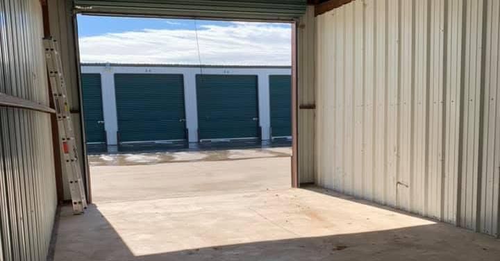 Open storage unit interior with concrete floor and rows of blue roll-up doors outside.