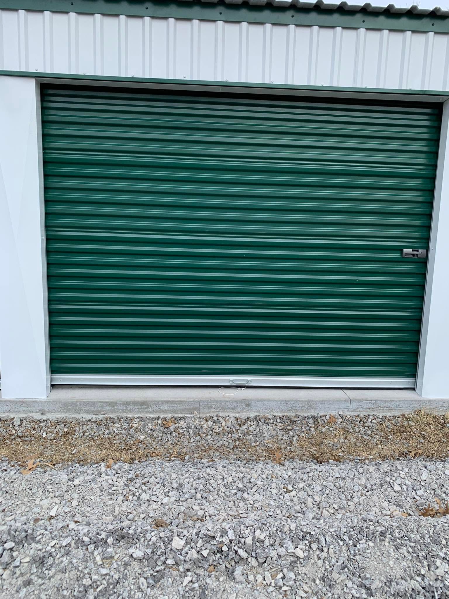 Green metal roll-up garage door on a white building with gravel in front.