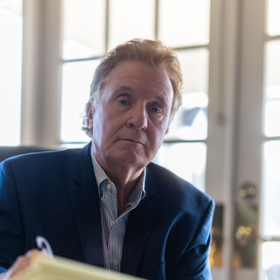 Man seated indoors near a bright window, wearing a dark blazer and striped shirt, looking toward the camera.