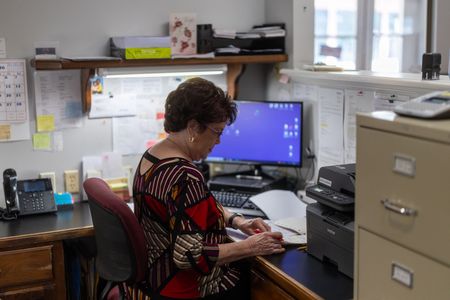 Person seated at an office desk, working on a computer in a cluttered cubicle.
