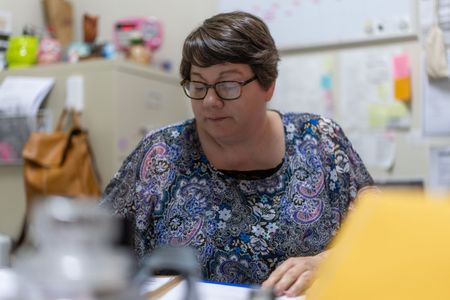 Woman wearing glasses working at a desk in a classroom or office setting