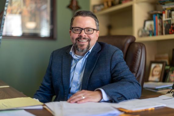 Smiling man in glasses sitting at a desk in an office, with papers and books around him.