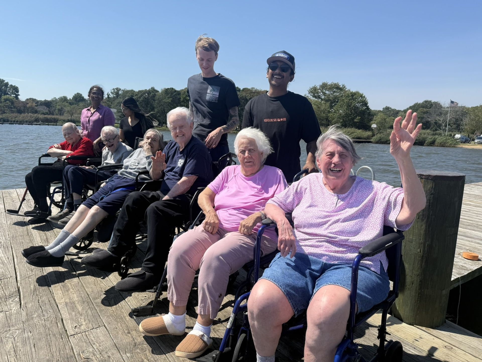 People seated on a dock by the water, smiling and waving on a sunny day.