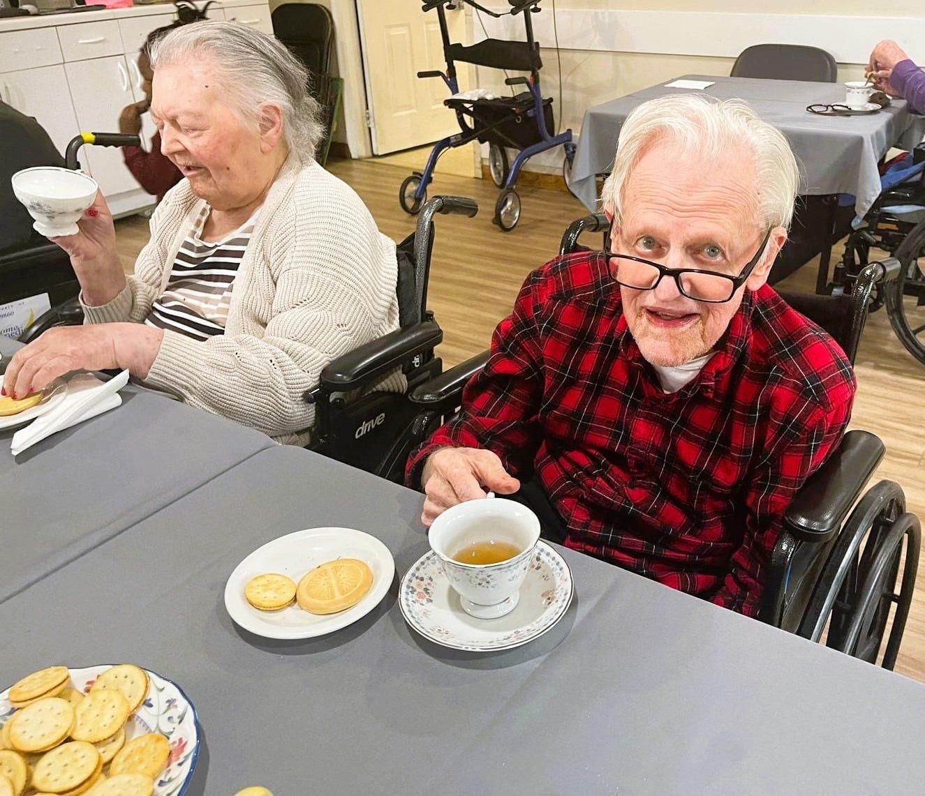 Two elderly women in wheelchairs enjoy tea and cookies at a table in a community room.