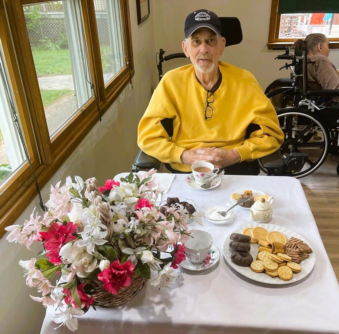 Elderly man in yellow sweater at a tea table with flowers, tea cups, and cookies by a window