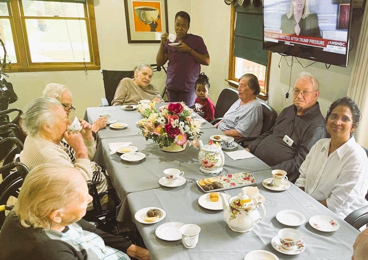 People seated around a dining table in a bright room, with tea cups, plates, and a flower centerpiece