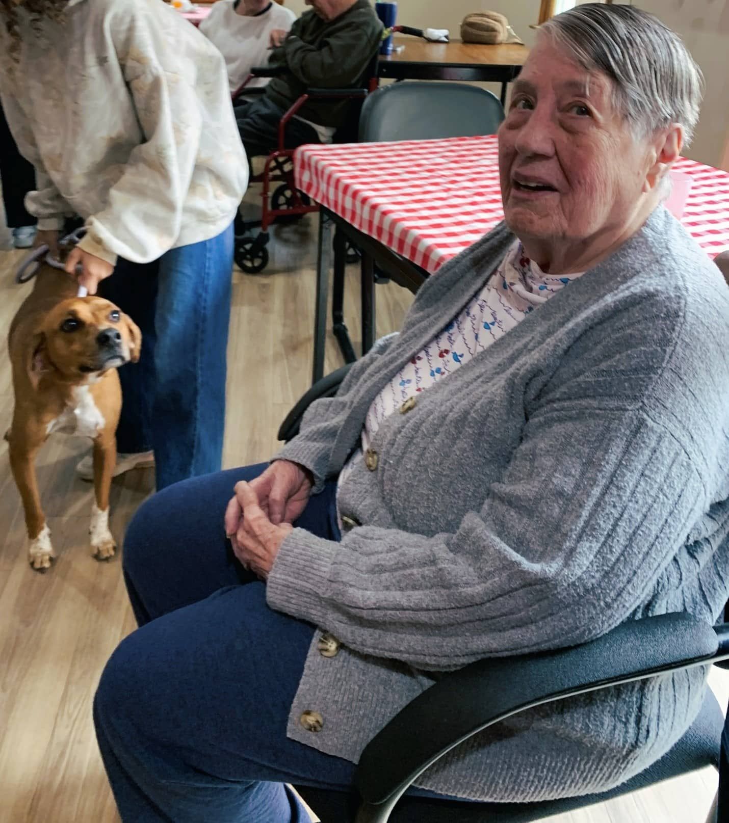 Woman sitting indoors in a chair, smiling, with a dog nearby in a dining hall