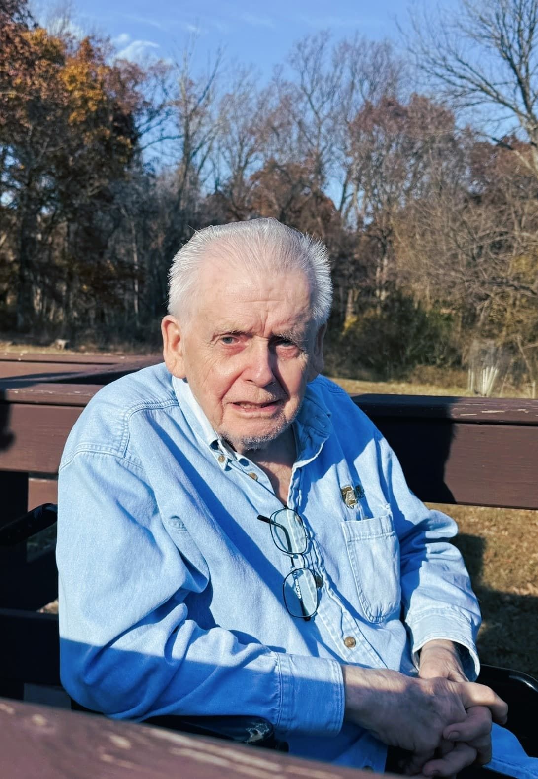Elderly man in a blue shirt sitting outdoors on a bench with trees in the background