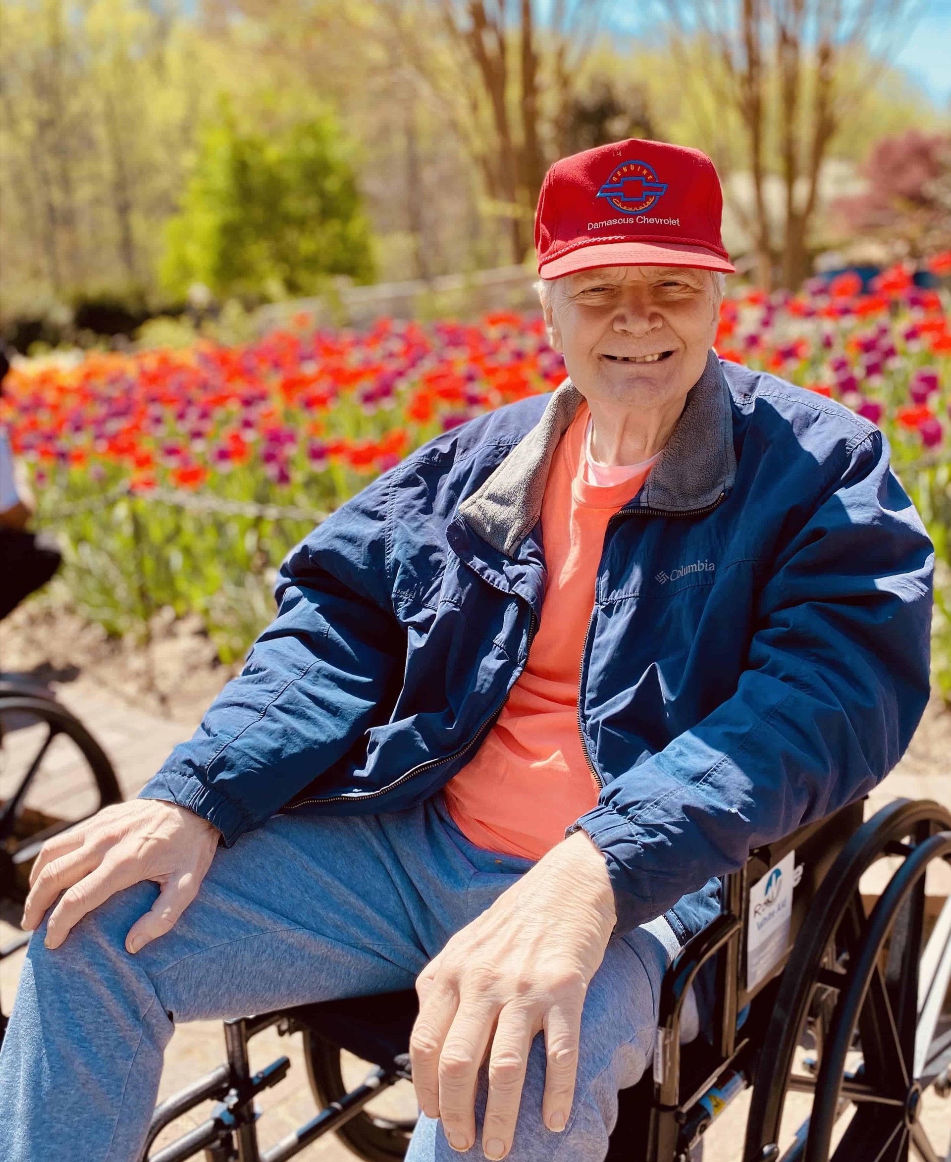 Person in a wheelchair wearing a red cap and blue jacket, sitting outdoors in a flower garden.
