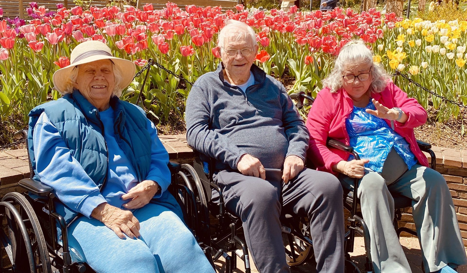 Three people sitting on a bench in front of blooming red flowers, smiling in the sun.