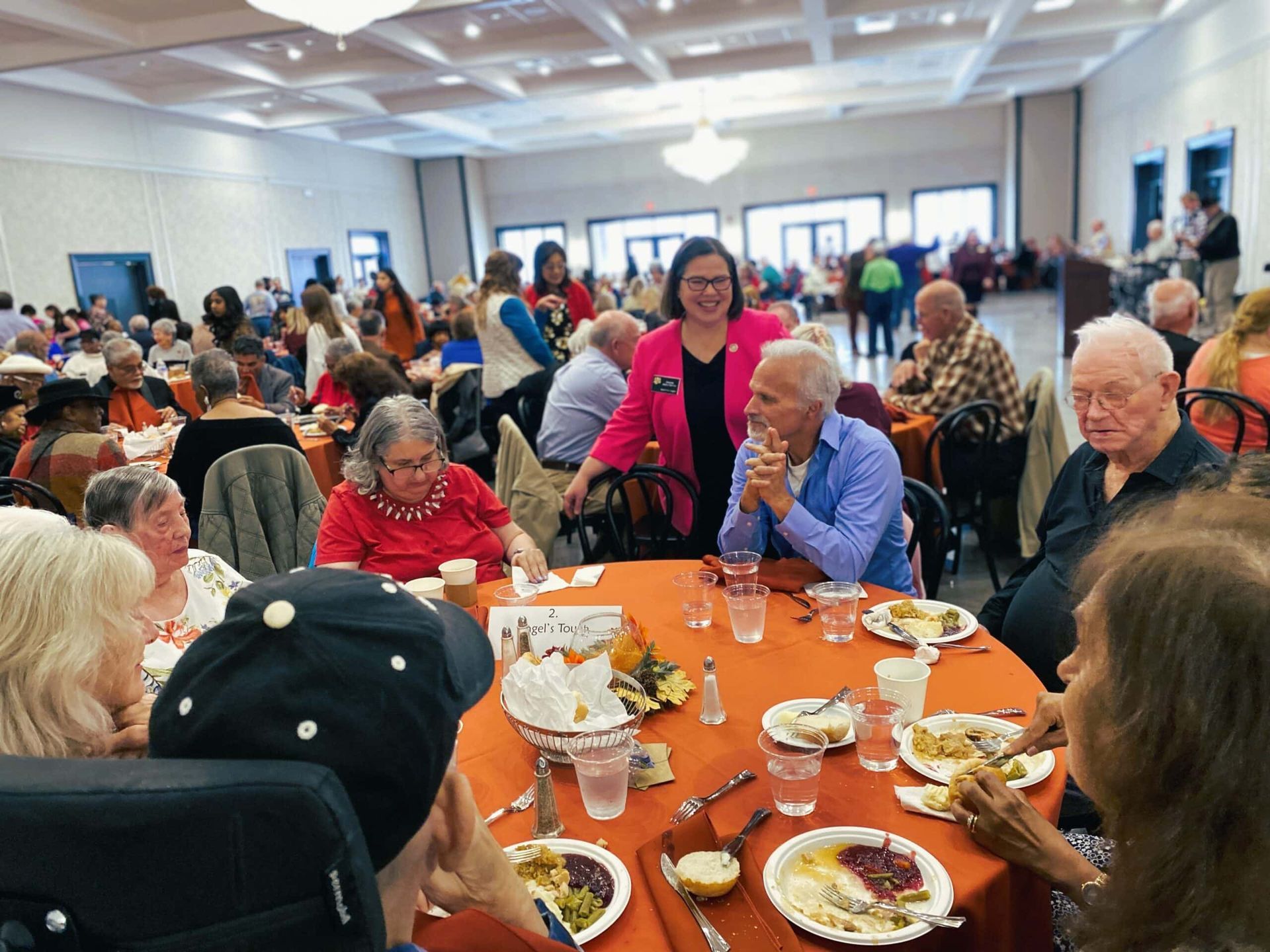 People seated around orange-clothed tables at a lively indoor banquet, with a woman standing and smiling nearby