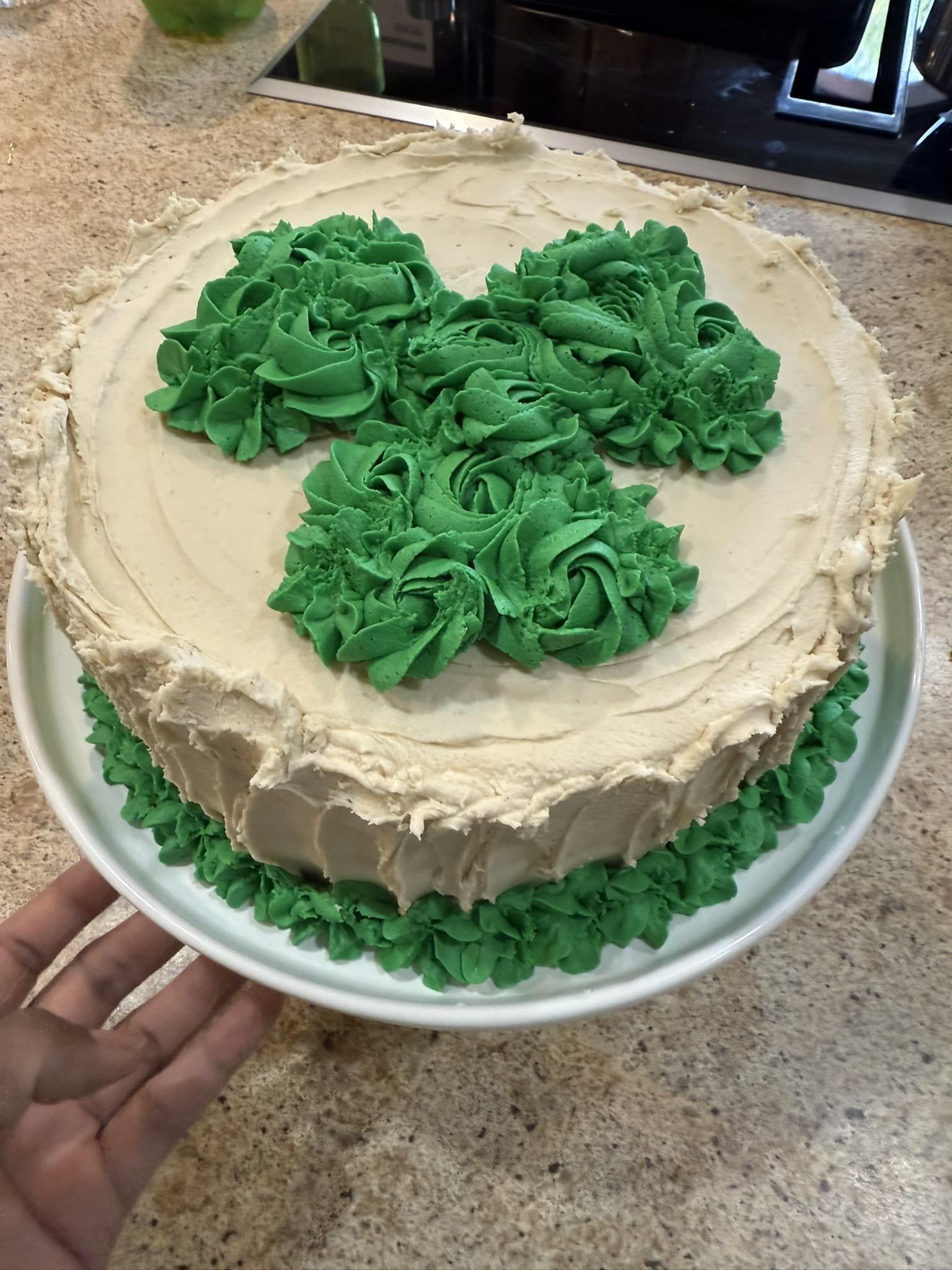 Round frosted cake with green rosettes on top and sides, on a white plate in a kitchen.