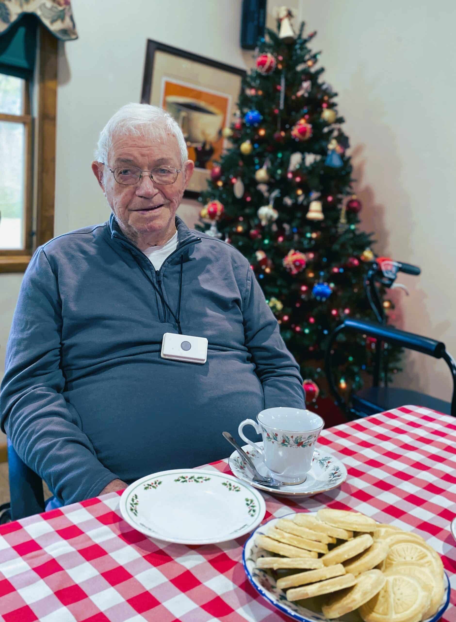 Man sitting at a table with tea and cookies beside a decorated Christmas tree.