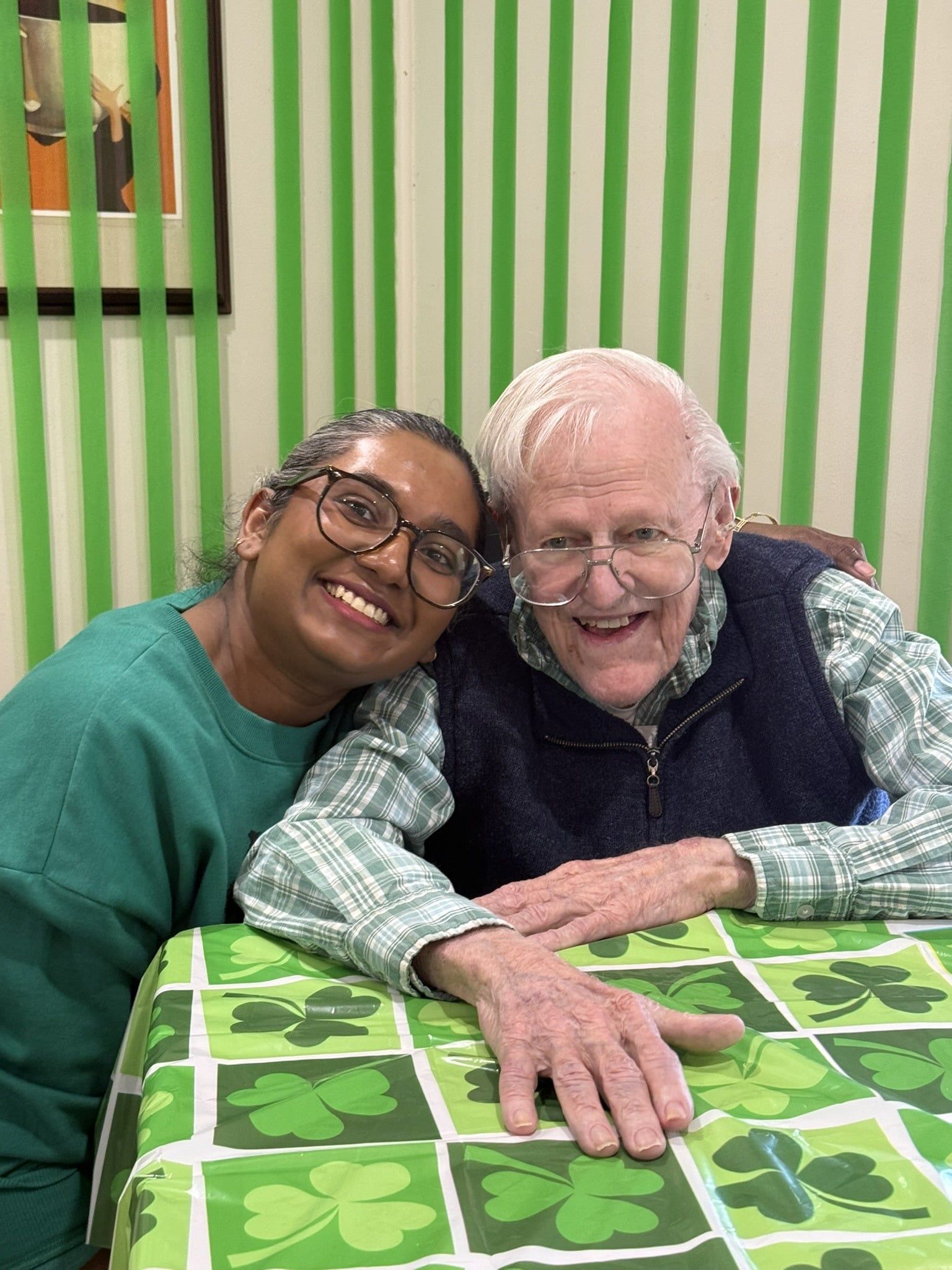 Two smiling people lean together at a green clover-covered table in a striped room.