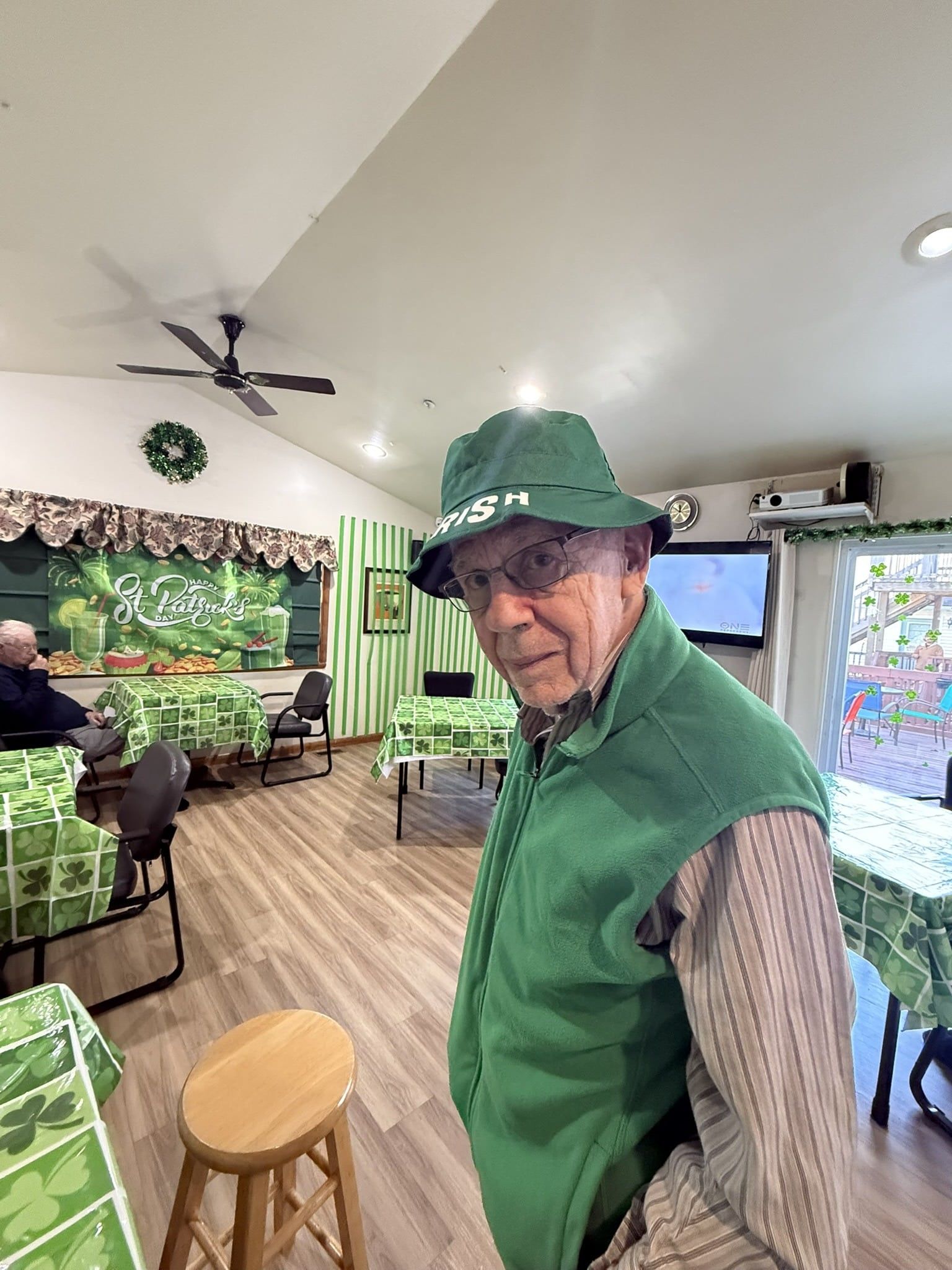 Man in green jacket and hat standing in a cozy café with wood floors and plants.