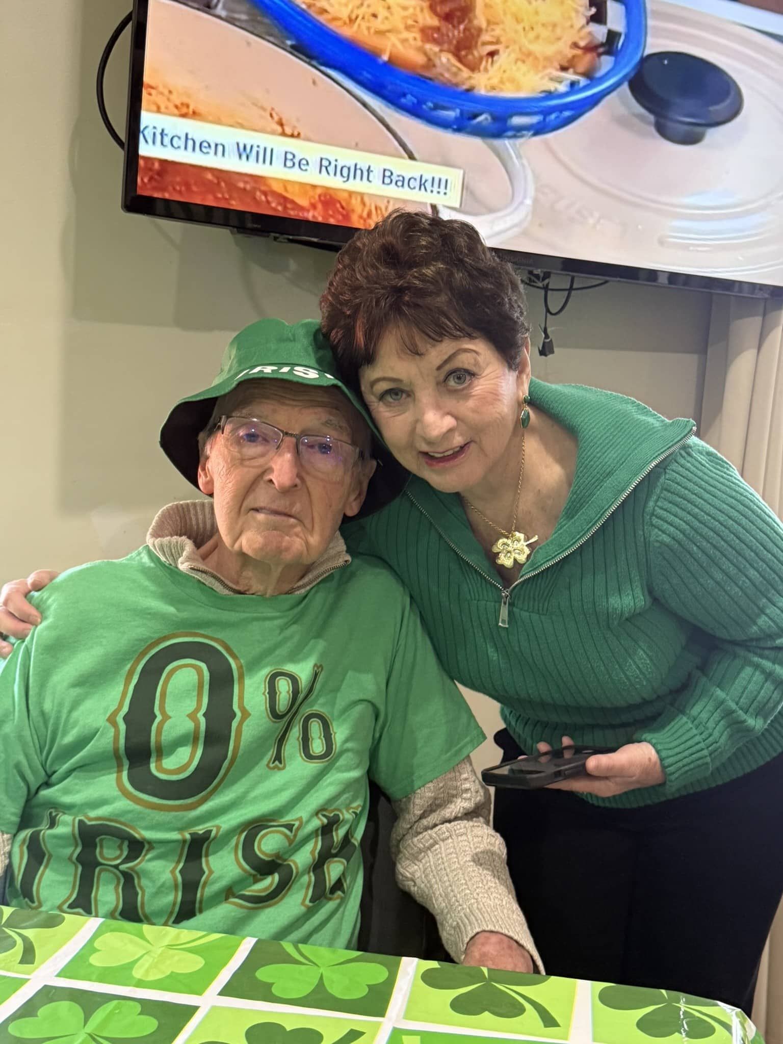 Two people in green shirts smiling together at a table in front of a TV screen