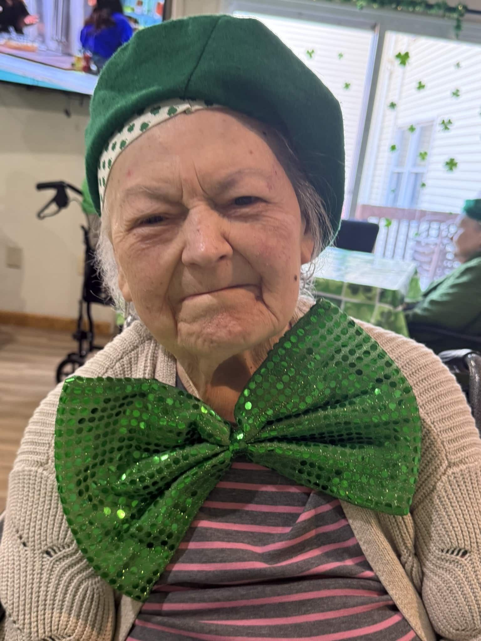 Elderly woman in green hat and oversized glittery bow, sitting indoors and smiling softly