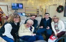 Five people smiling on a couch in a decorated living room with warm holiday lights and a wreath.