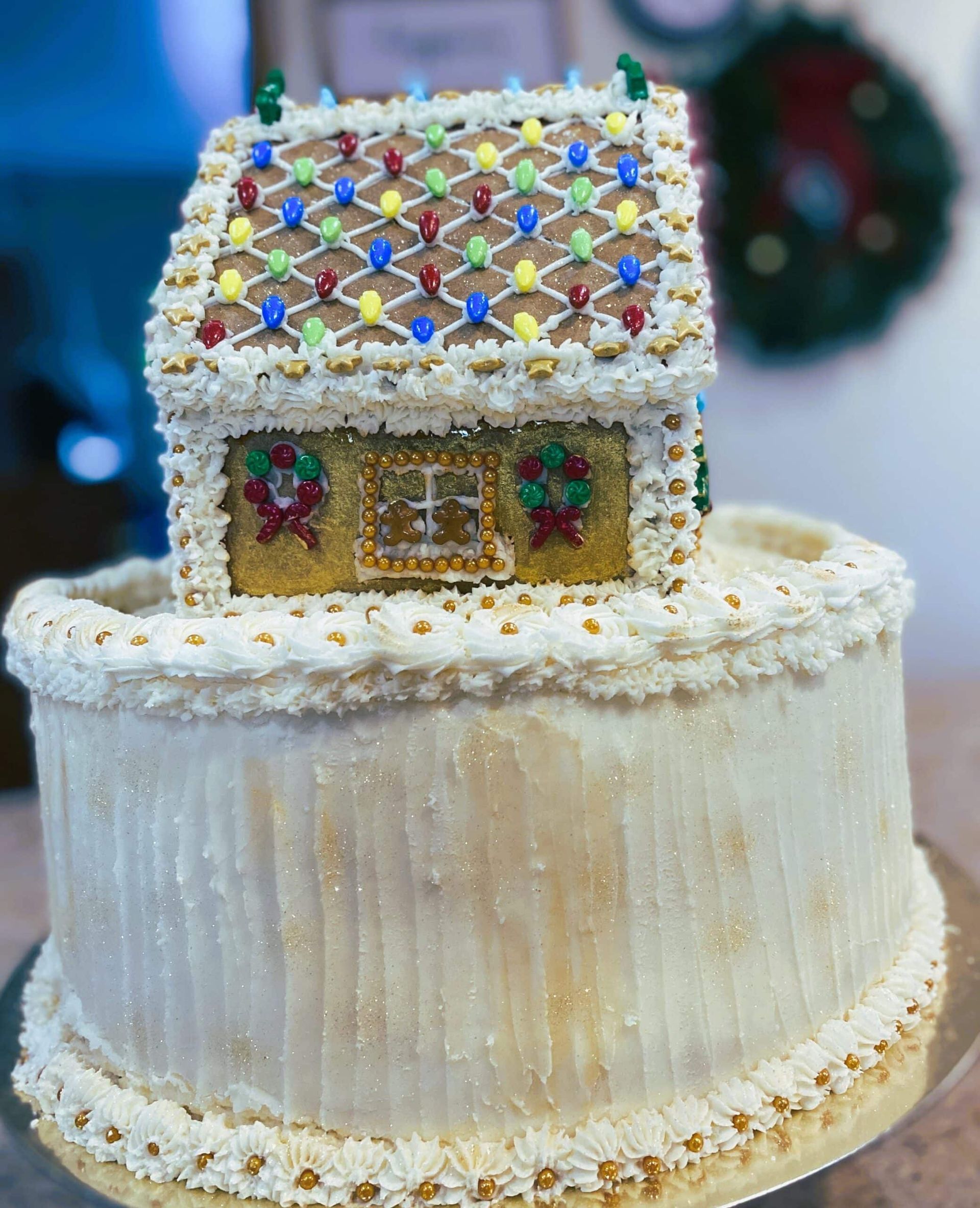 Two-tier white frosted cake with a small decorated house topper, covered in colorful candy pieces