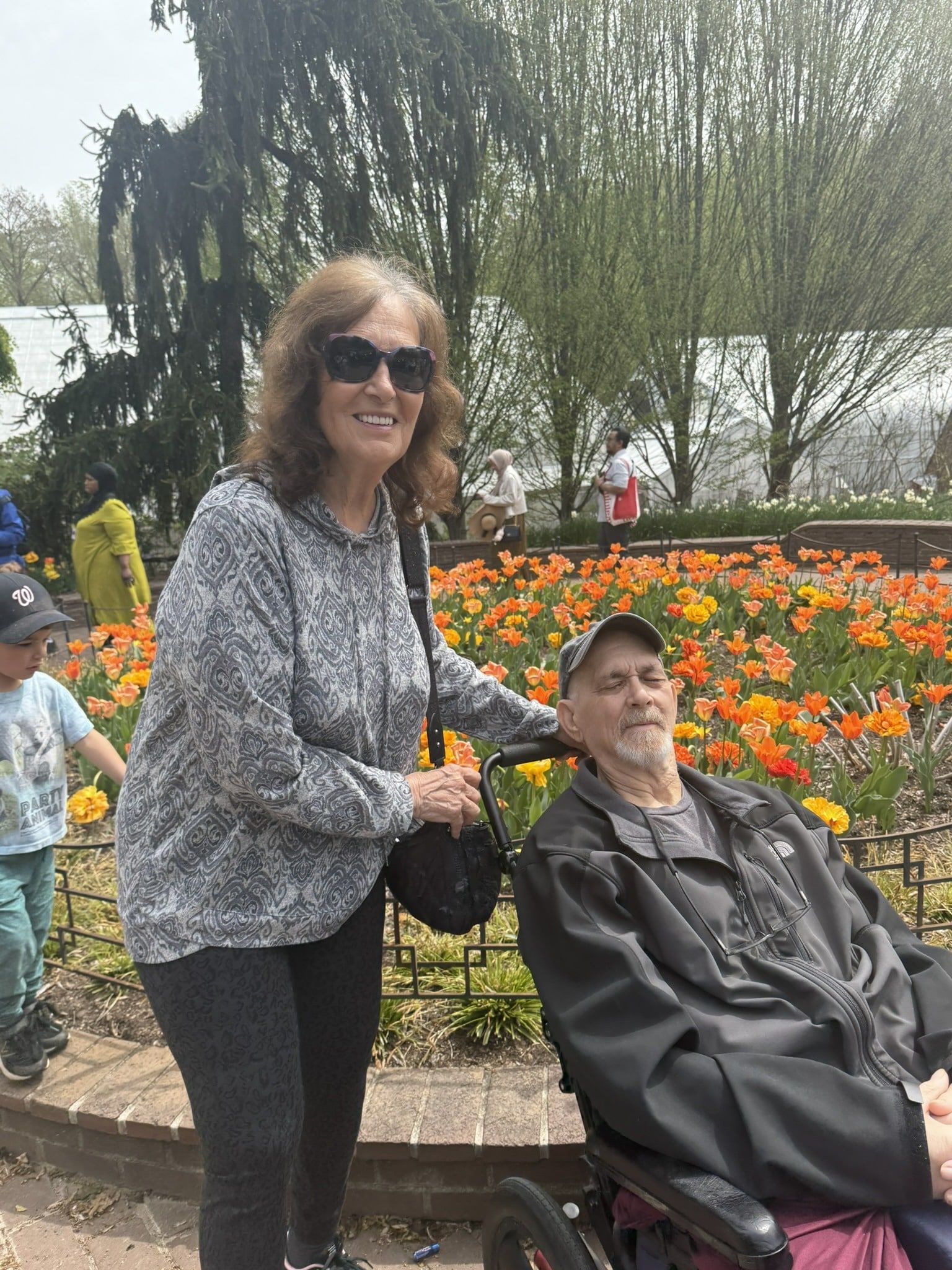 Woman standing beside a seated person in a wheelchair near a colorful flower bed in a park