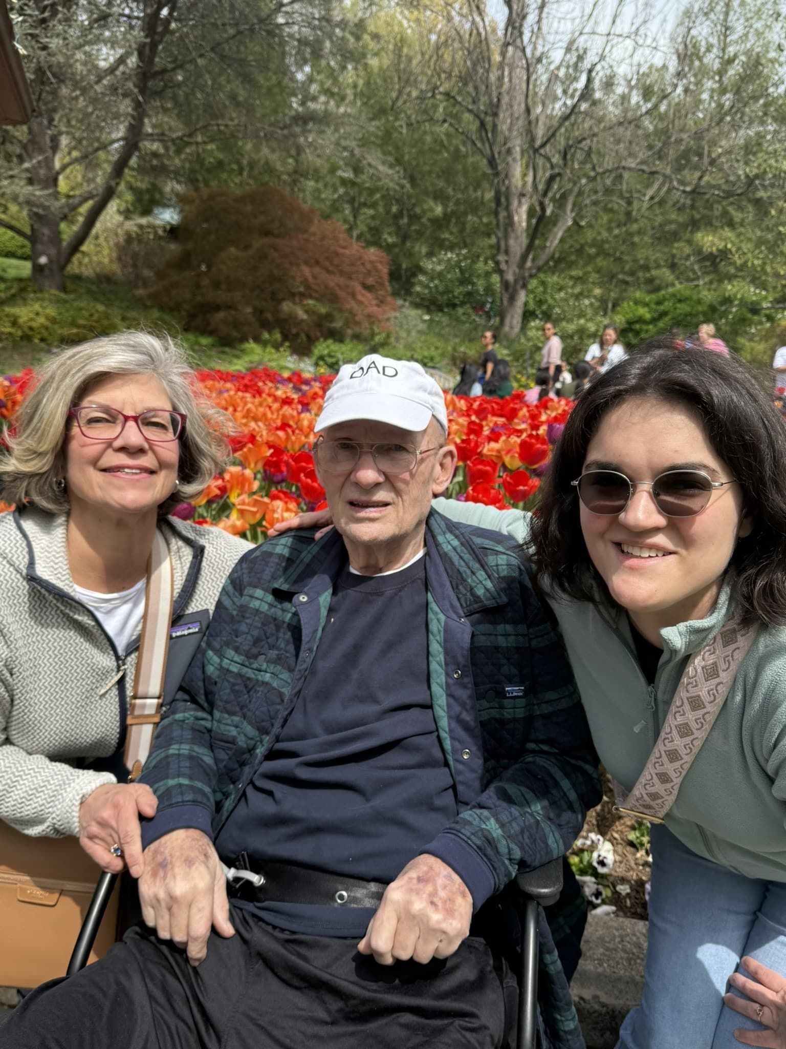 Three people seated outdoors in a garden, with bright red flowers and trees behind them.