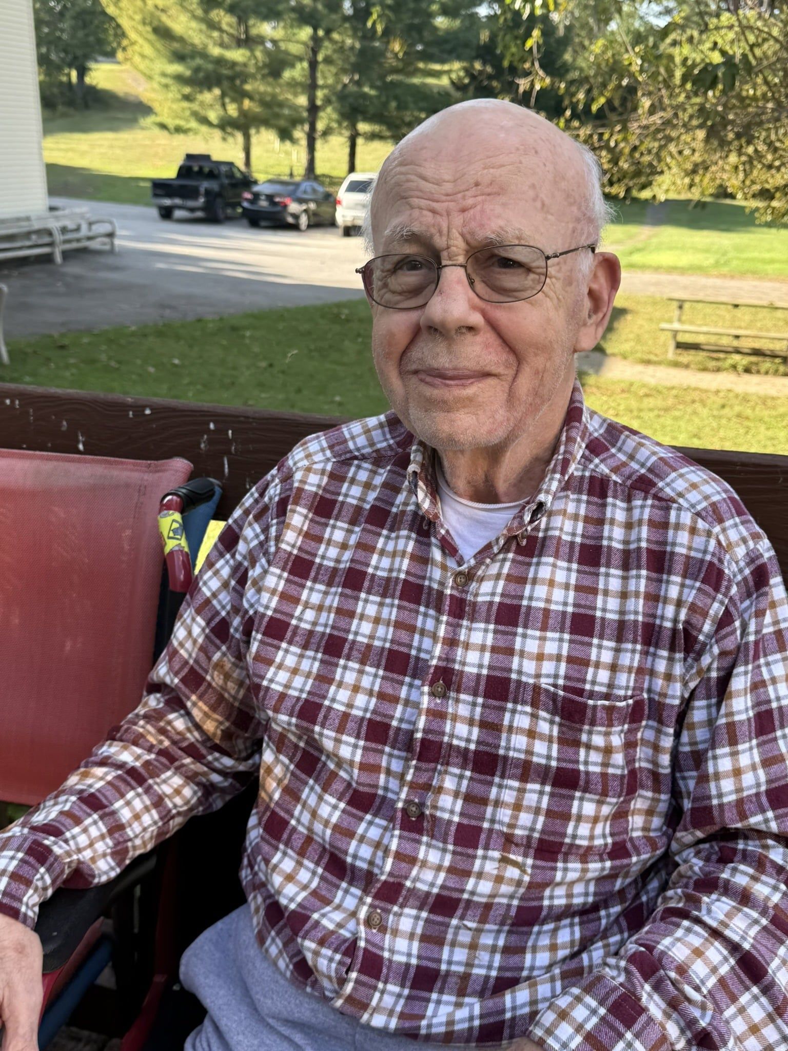 Elderly man in a red plaid shirt sitting outdoors on a porch, smiling at the camera.