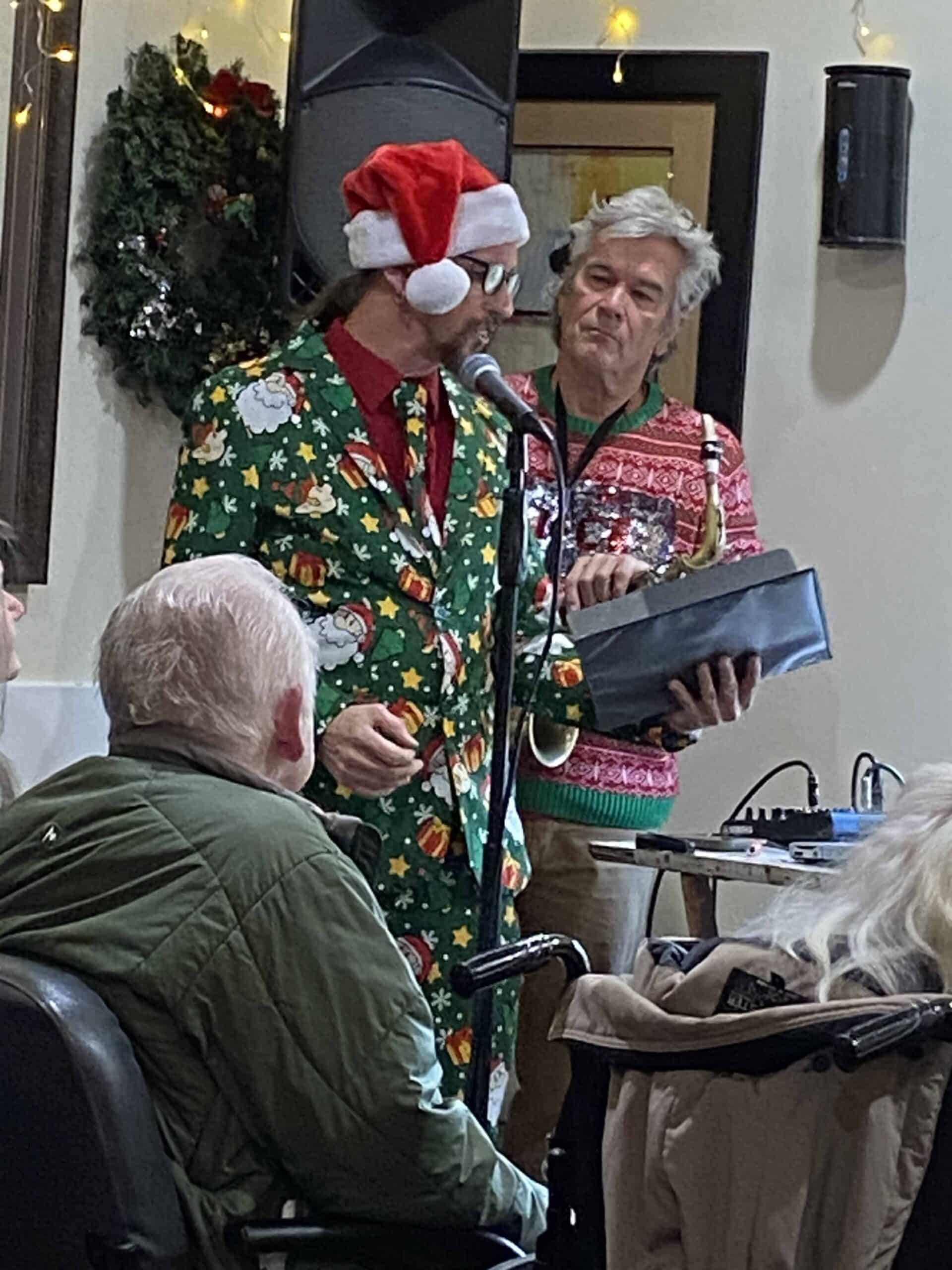Two people in festive sweaters and Santa hats stand indoors holding a tablet near a decorated holiday wreath.