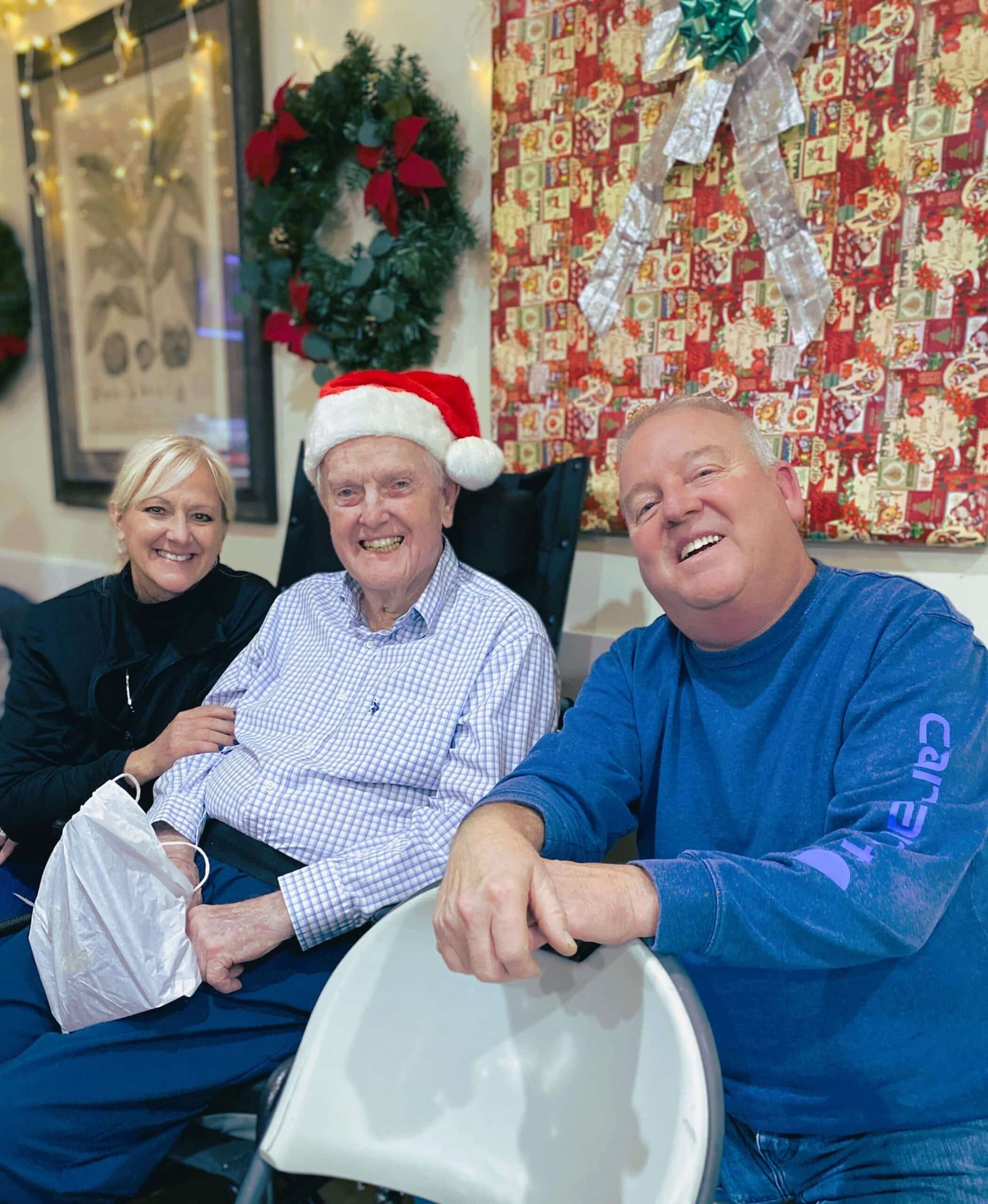 Three people seated indoors, smiling in front of festive holiday decorations; one wears a Santa hat.