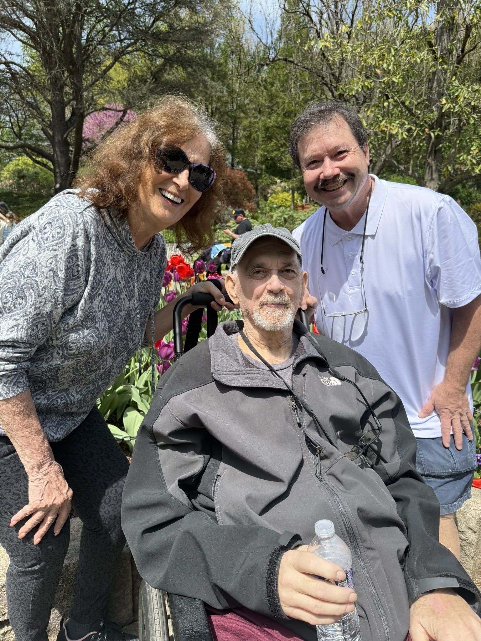 Three smiling people outdoors, one seated in a wheelchair, with flowers and trees in the background.