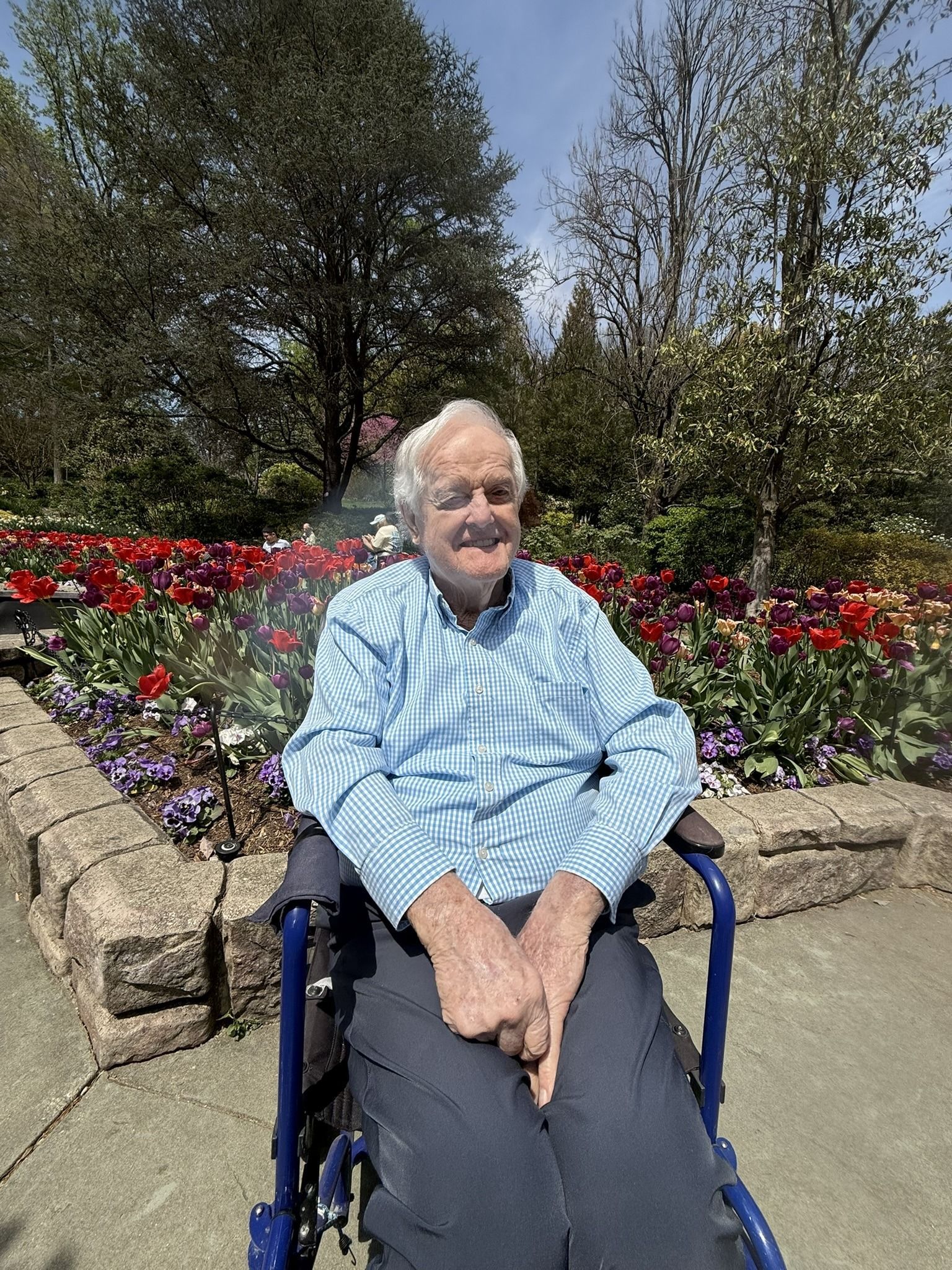 Person seated in a wheelchair in a flower garden, wearing a light blue shirt and smiling at the camera.