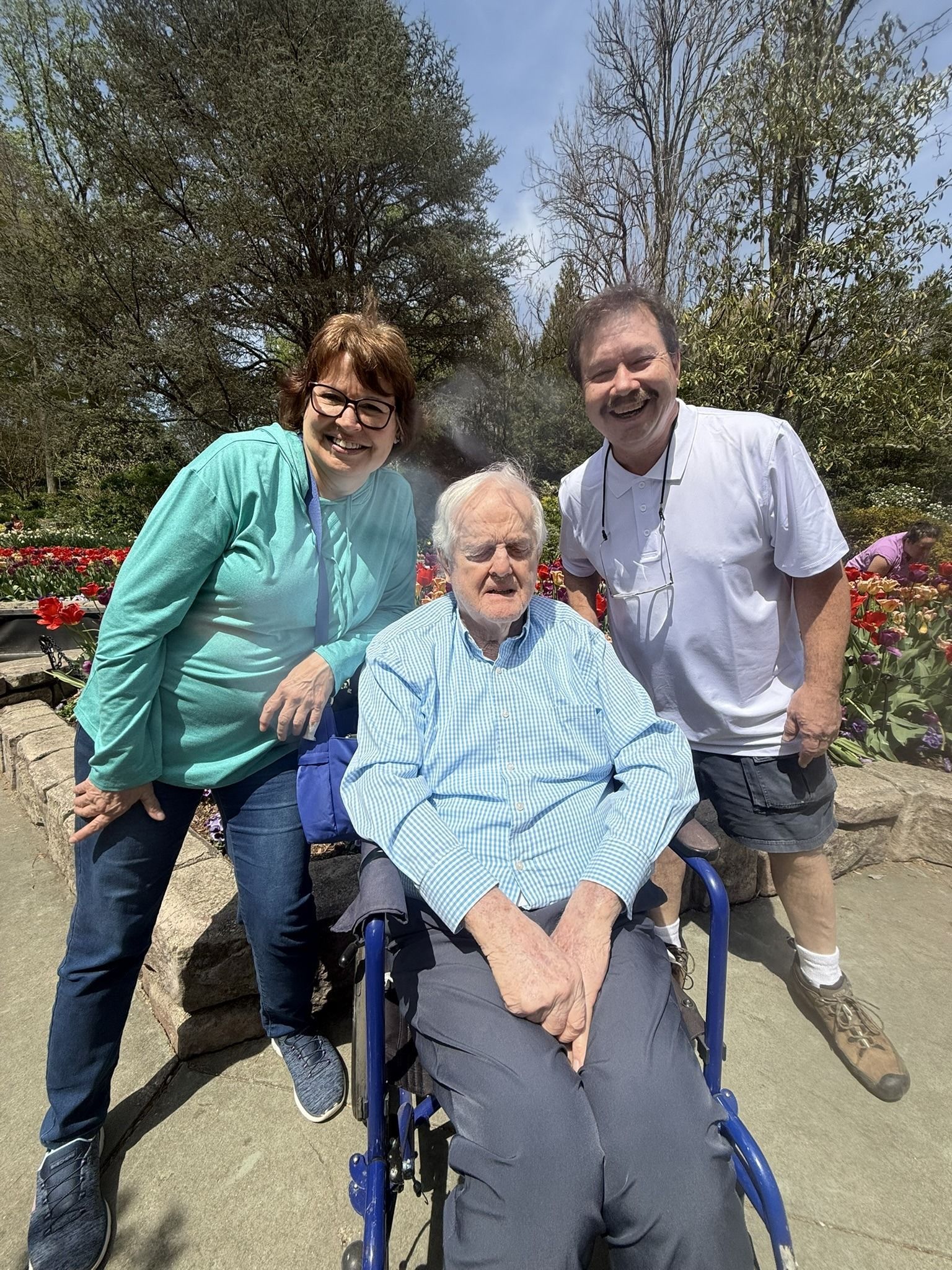 Three people smiling outdoors, one seated in a wheelchair in a sunny garden