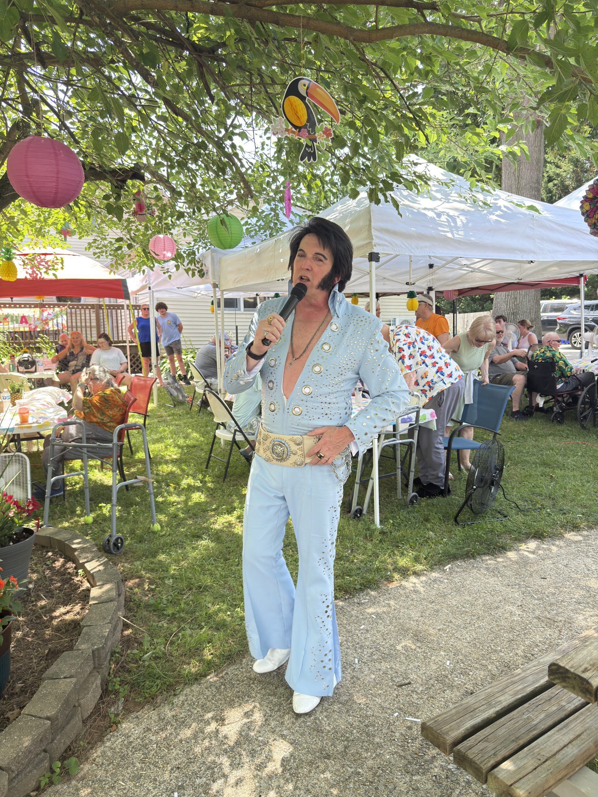 Person singing into a microphone at an outdoor market, wearing a floral top and jeans under a tree.