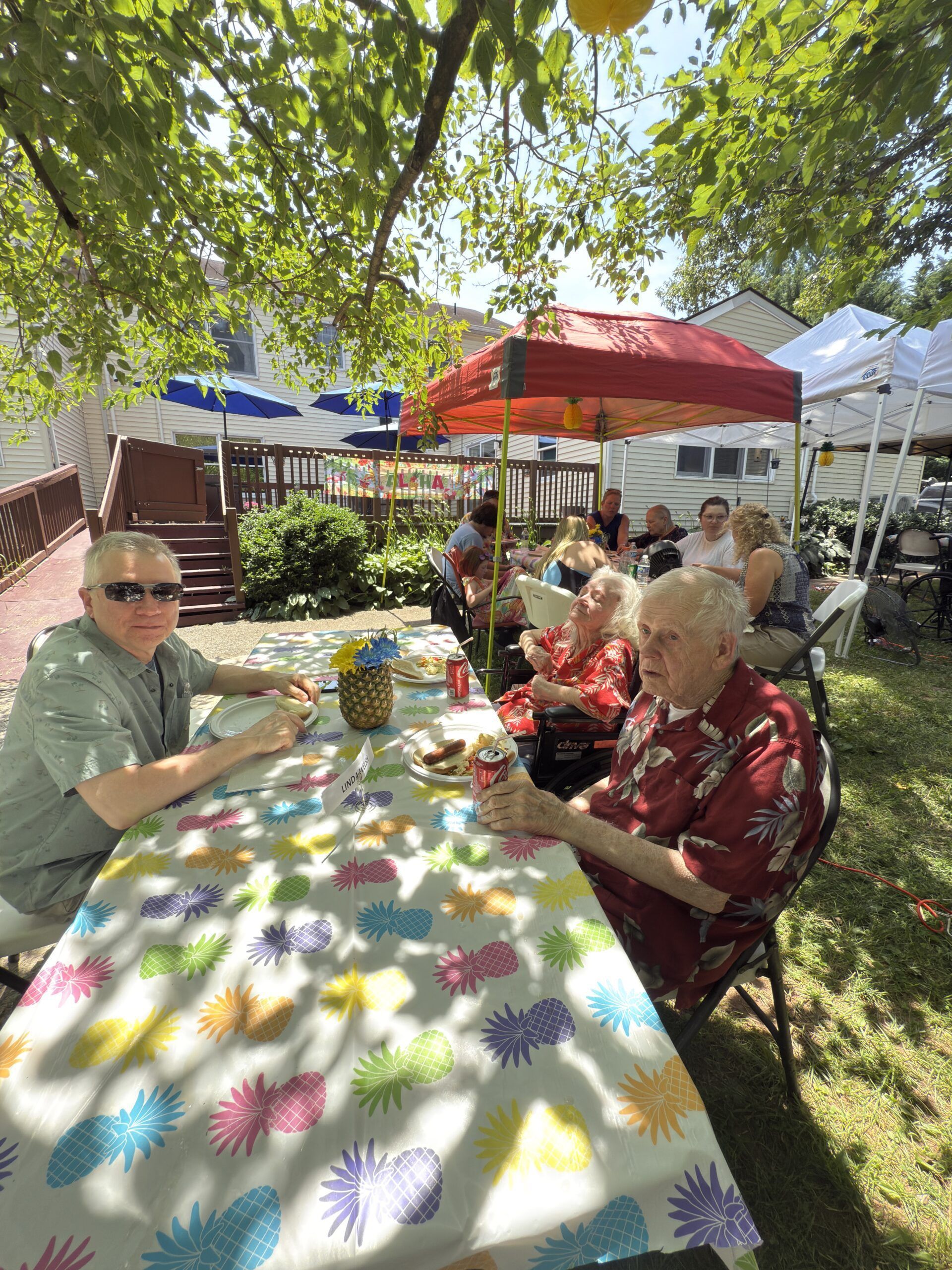 People seated at a colorful outdoor table under trees and umbrellas, enjoying a sunny garden gathering