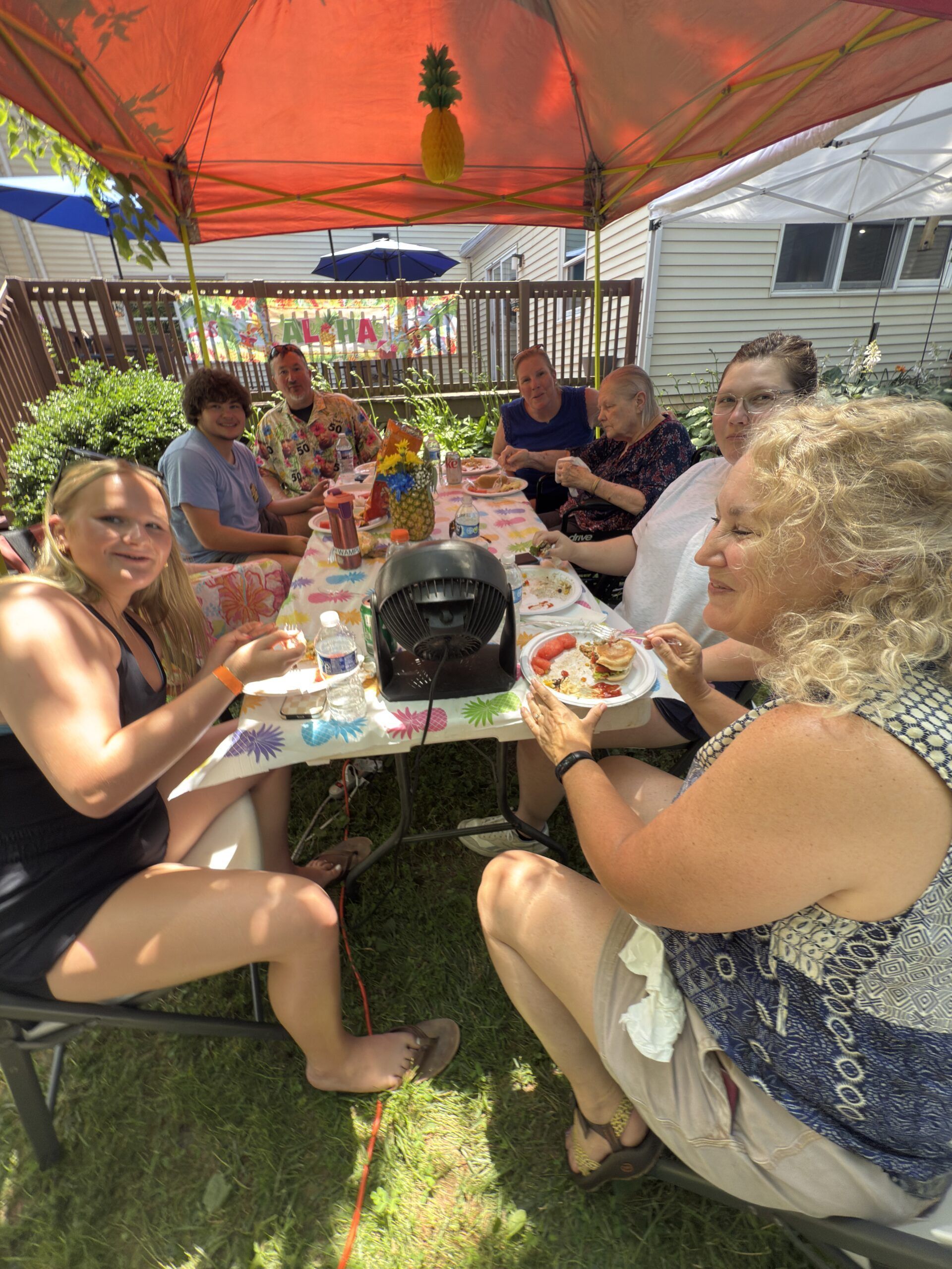 Friends eating at a picnic table under orange and blue canopies in a sunny backyard