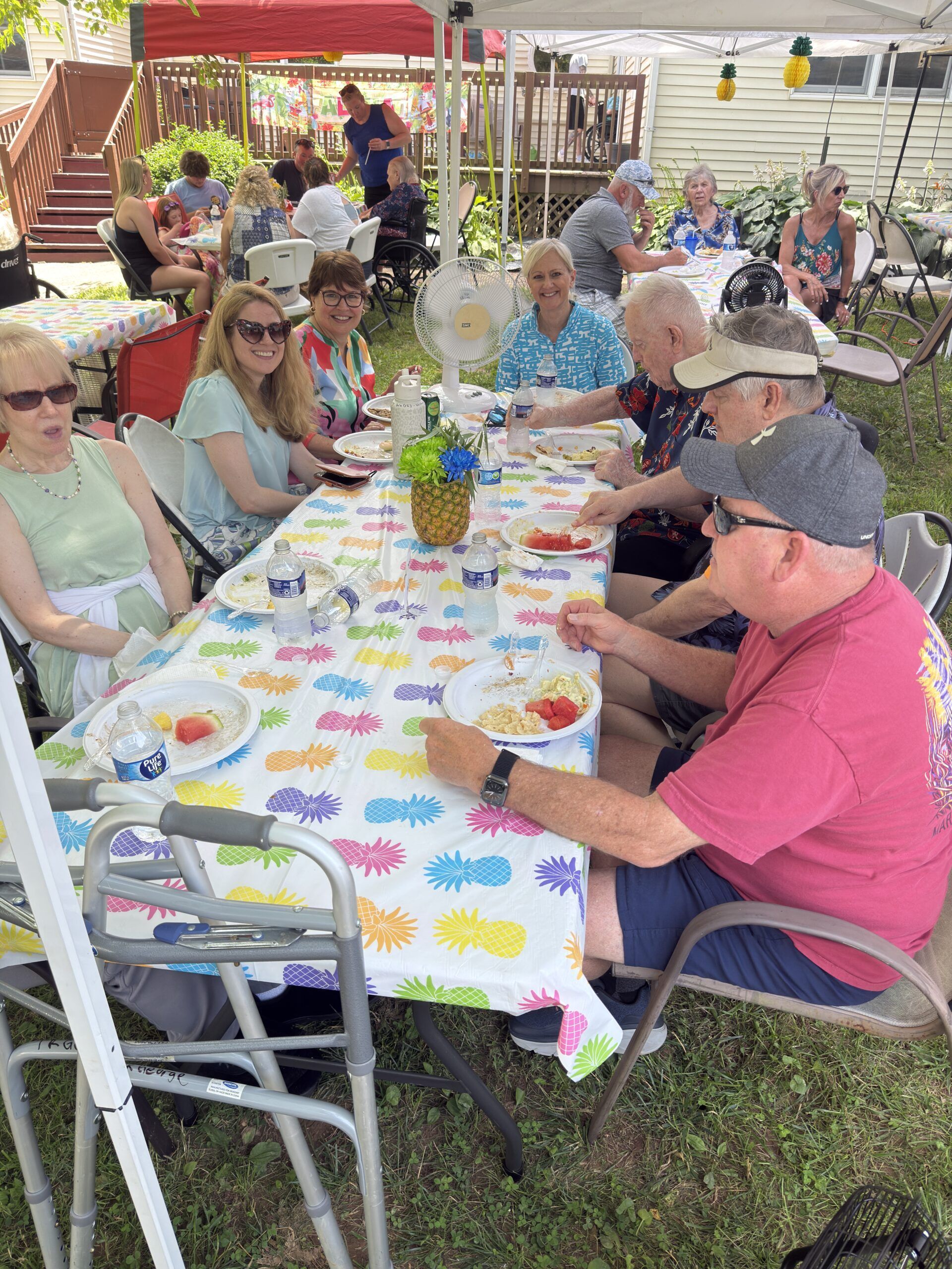 People gathered around a long table outdoors, sharing a meal under a canopy with colorful tablecloth and flowers.