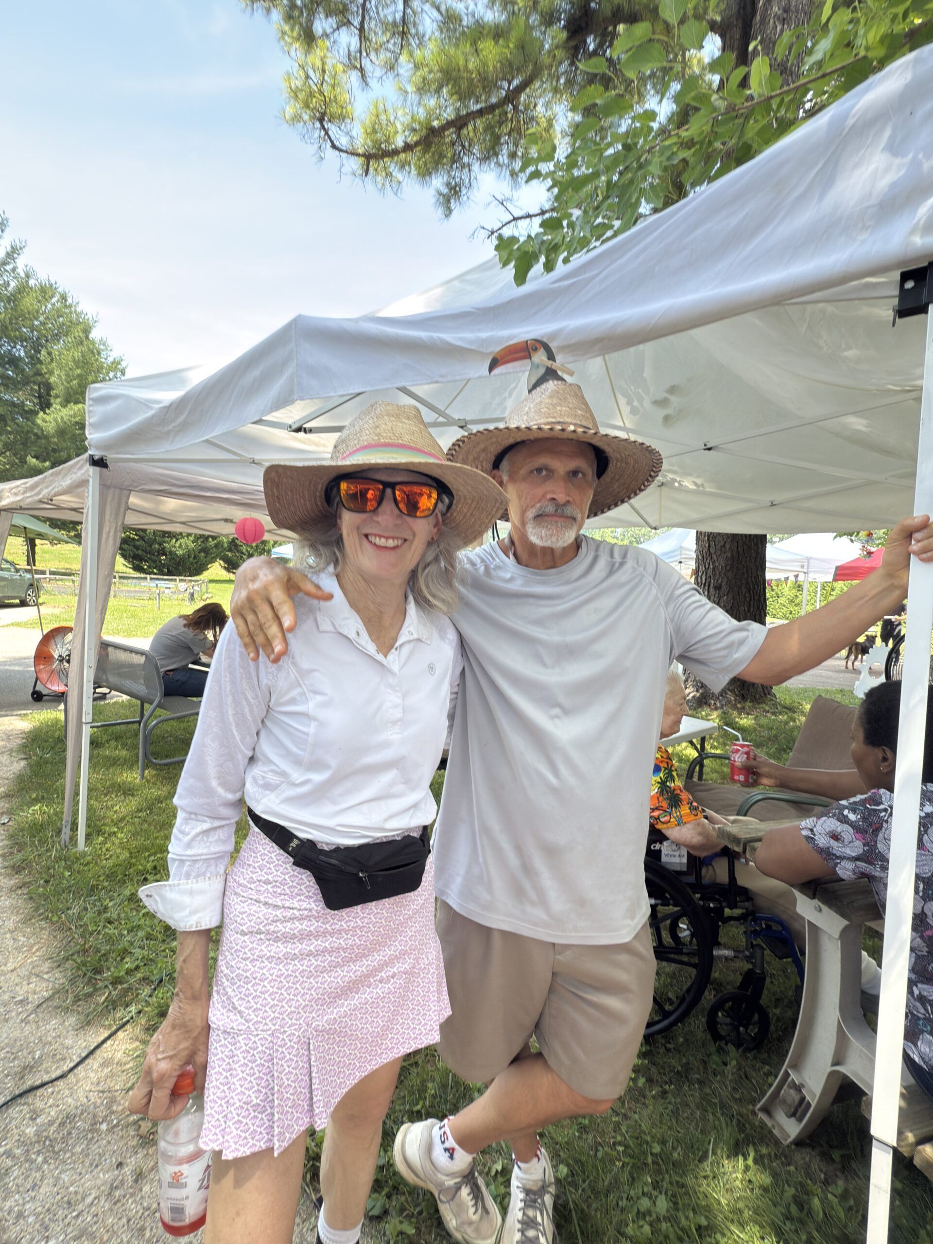 Two people smiling under a white canopy at an outdoor event, wearing hats and casual summer clothes.