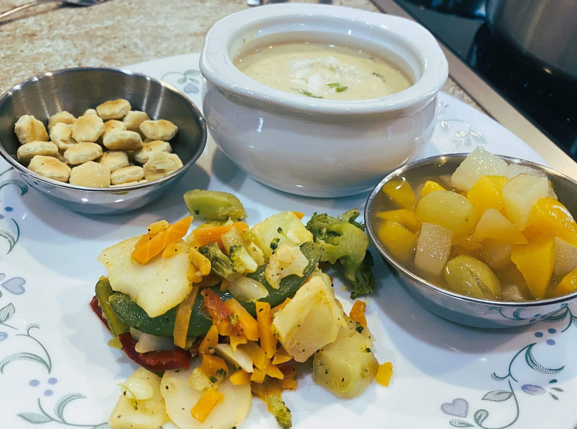 Three small bowls of snacks and soup on a floral tablecloth, with mixed vegetables in the center.