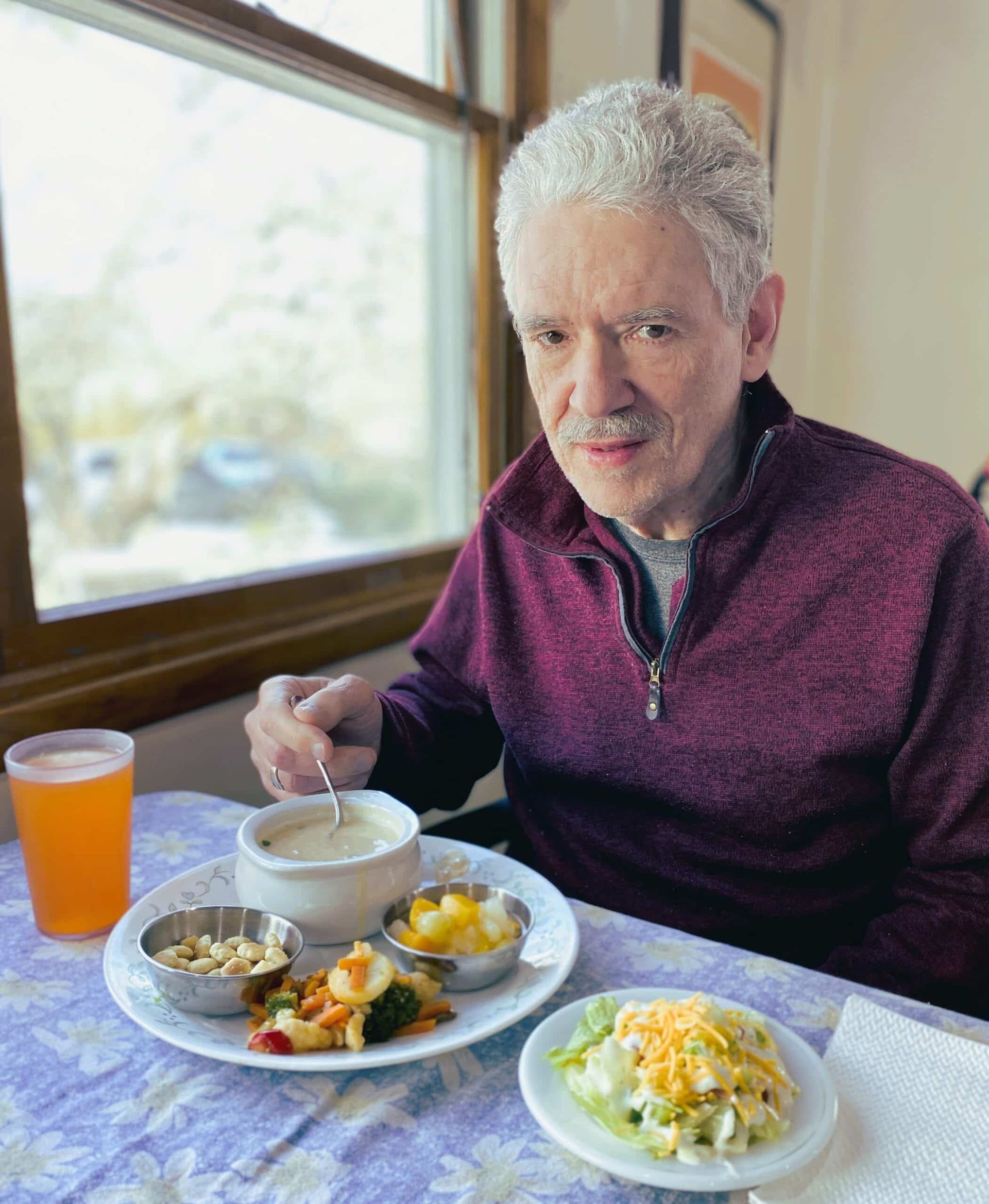 Man eating a meal at a table by a window, with orange drink and several dishes.
