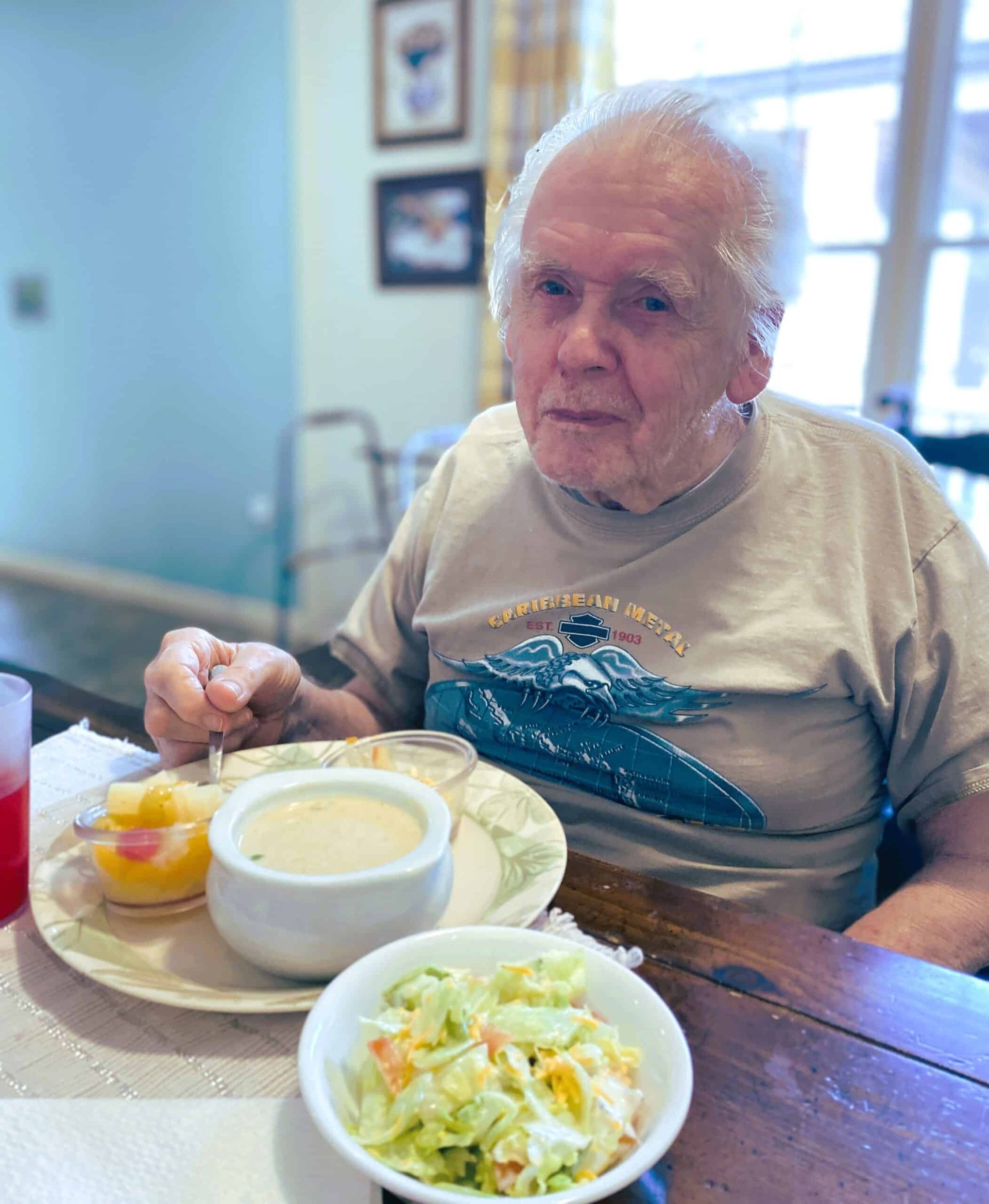 Elderly person at a table with soup, salad, and fruit, smiling in a bright dining room
