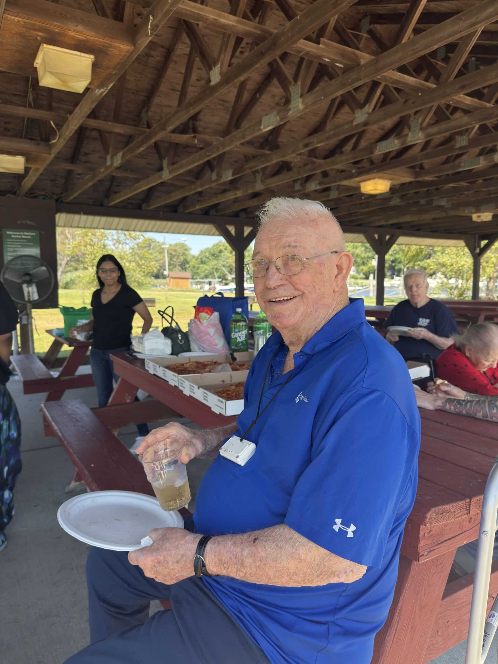 Smiling man in blue shirt holding a plate at a picnic shelter with benches and other people in the background