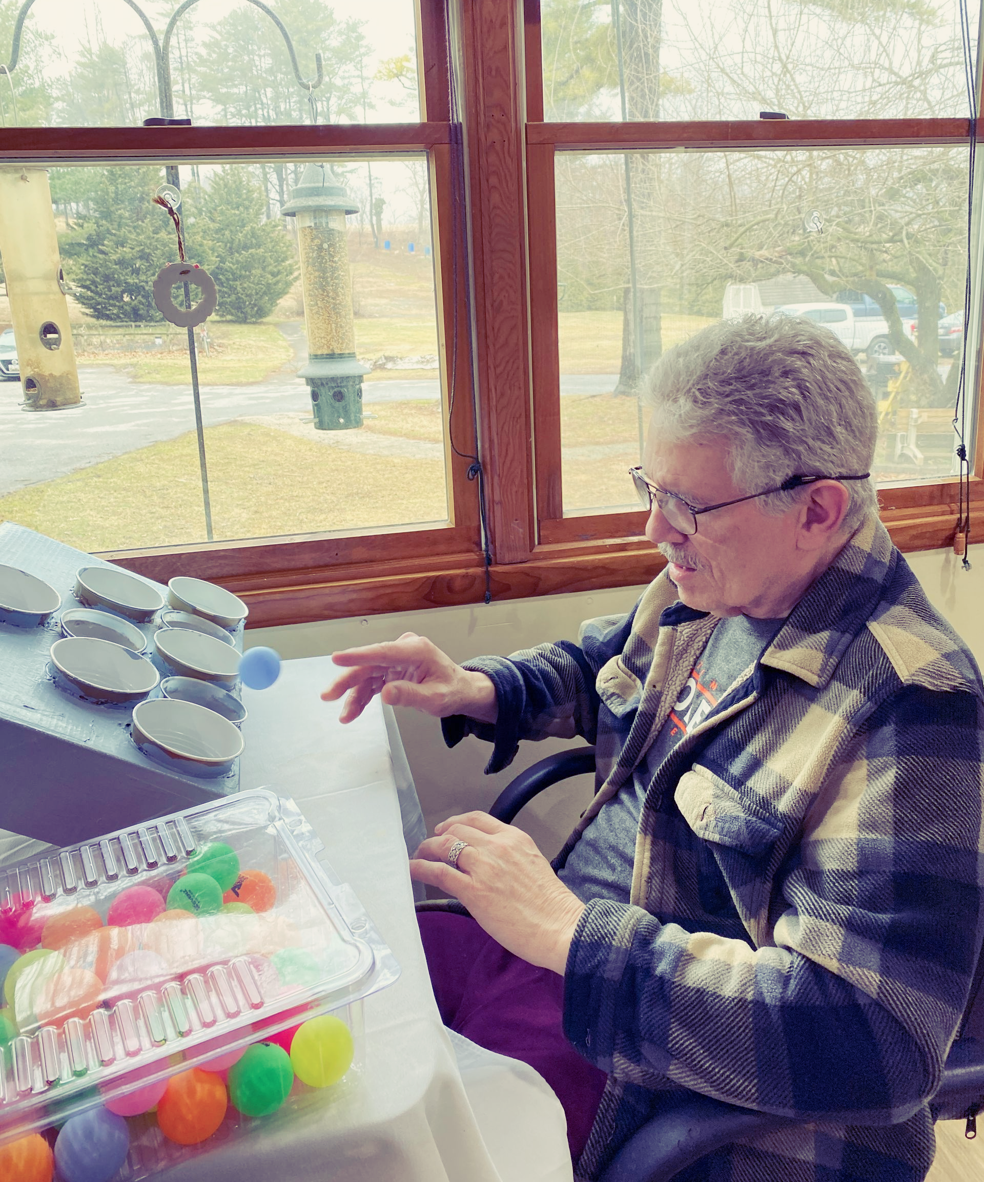 Elderly person sits by a window, reaching toward colorful balls on a table with a white tray and gray items.