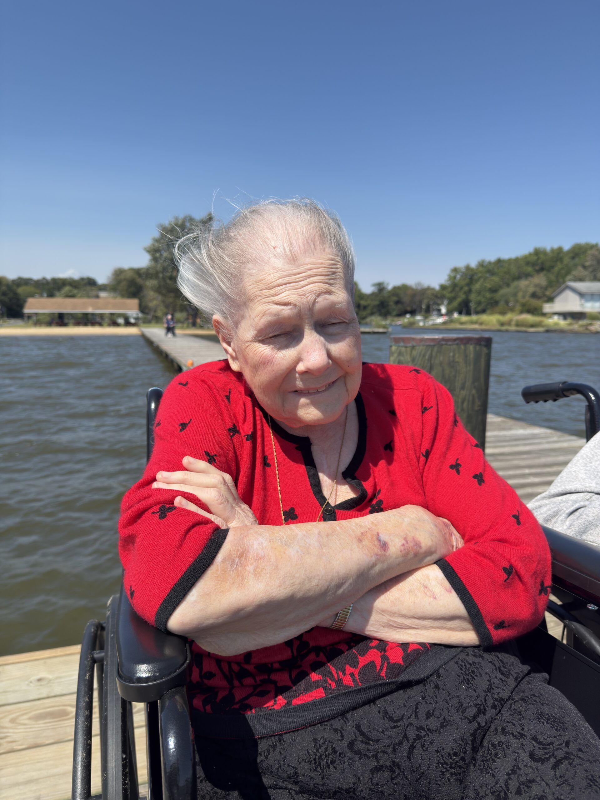 Elderly woman in a red sweater sits by the water, arms crossed, in a wheelchair on a sunny day.