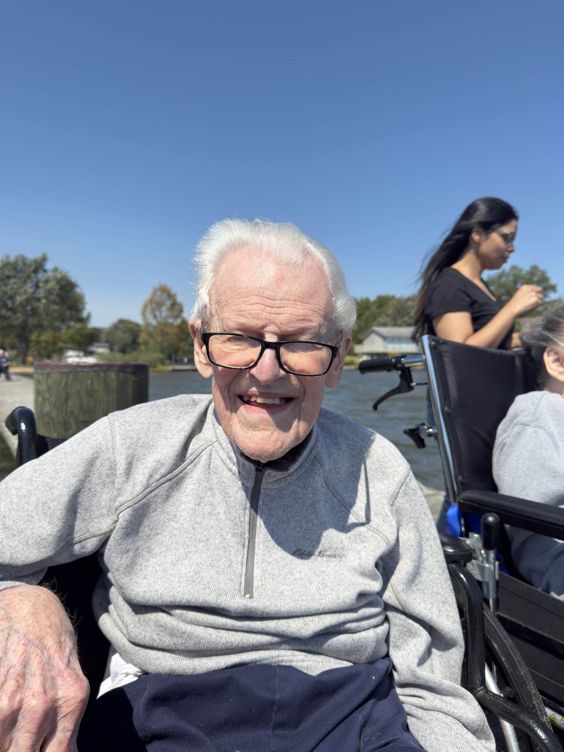 Smiling man in a gray sweatshirt sitting outdoors on a sunny day, with another person in the background.