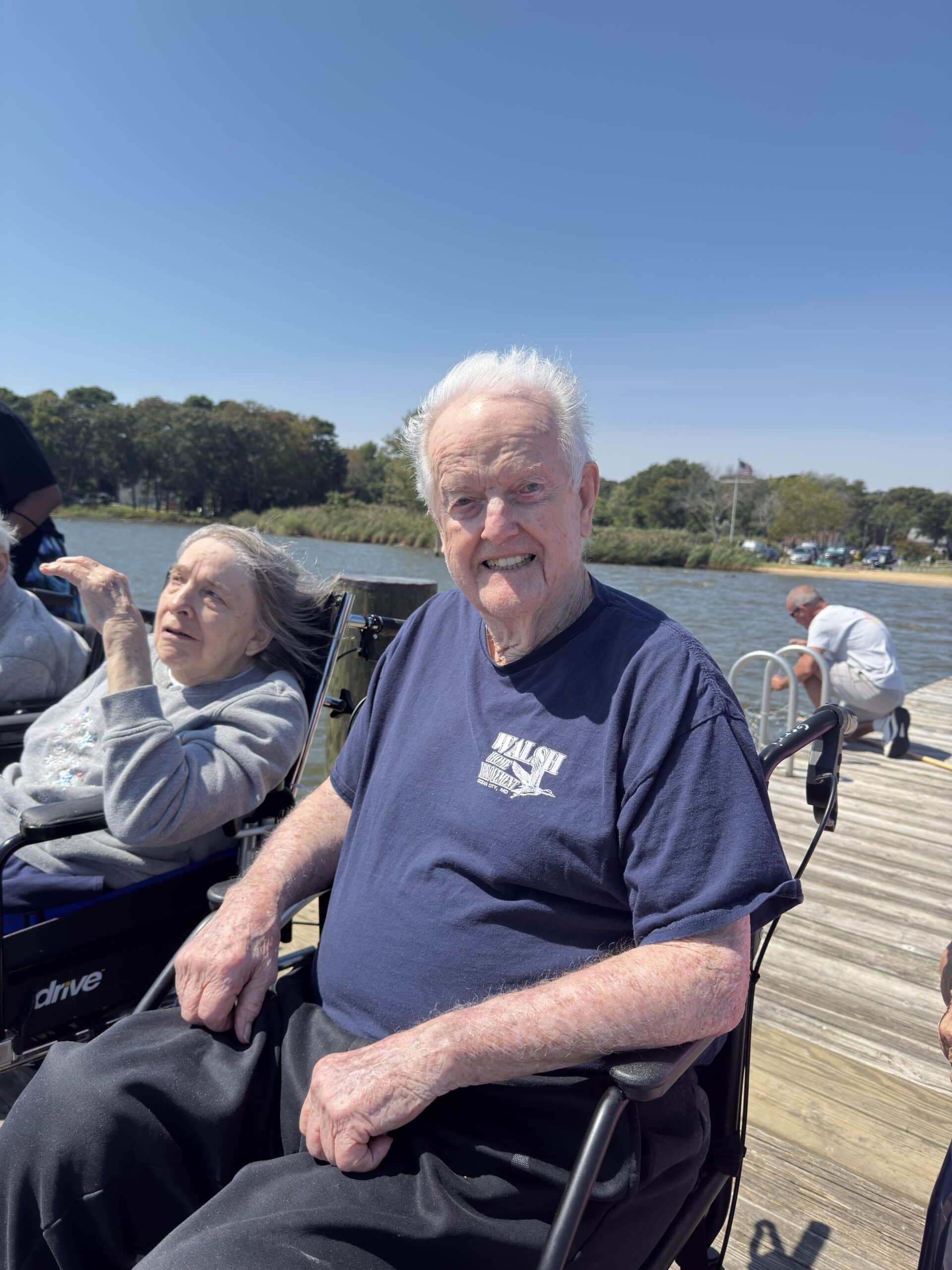 Smiling man in a navy T-shirt sits in a wheelchair outdoors by the water on a sunny day