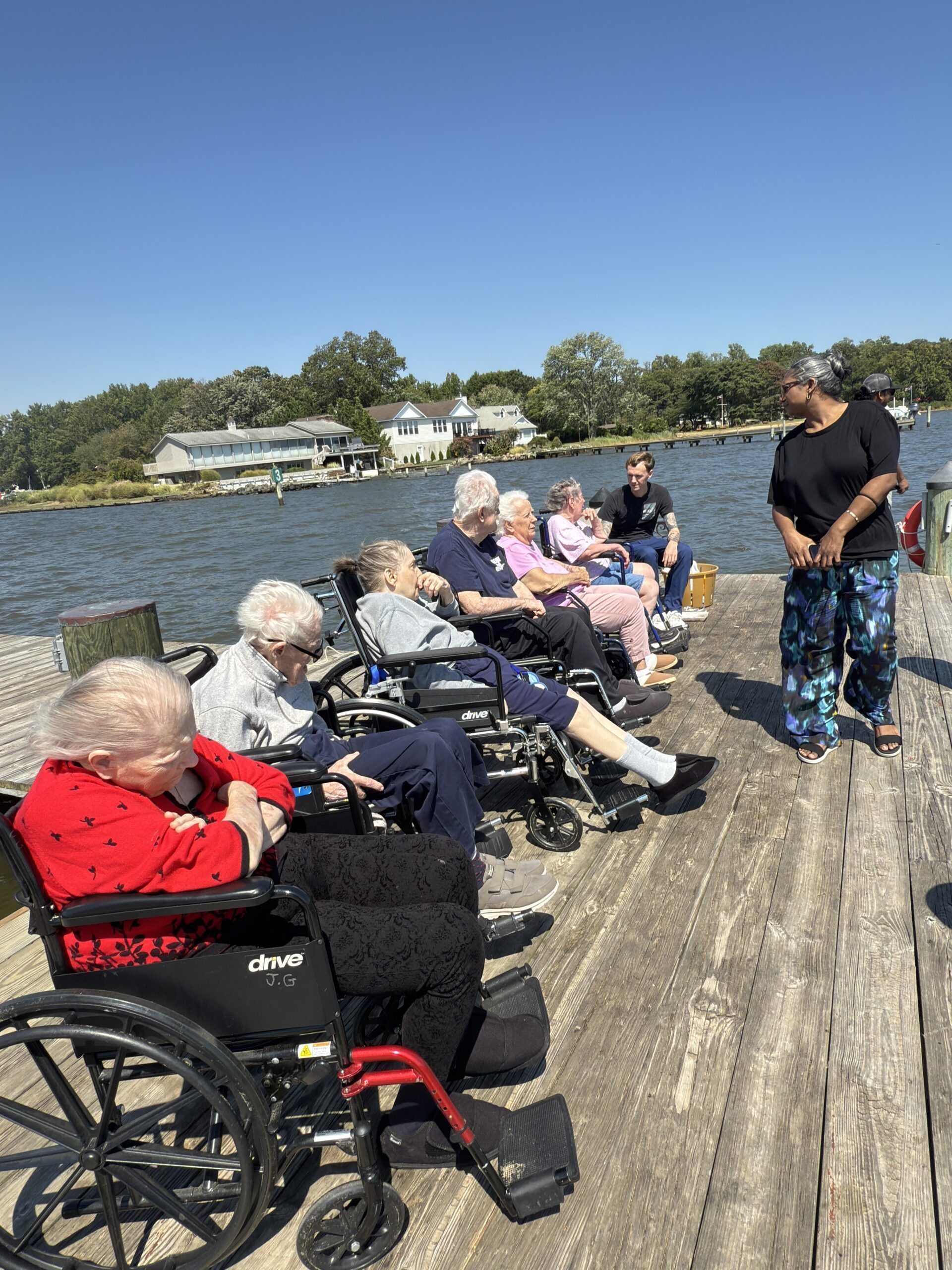 Group of people in wheelchairs on a wooden dock by the water, with a man standing nearby under a clear sky.