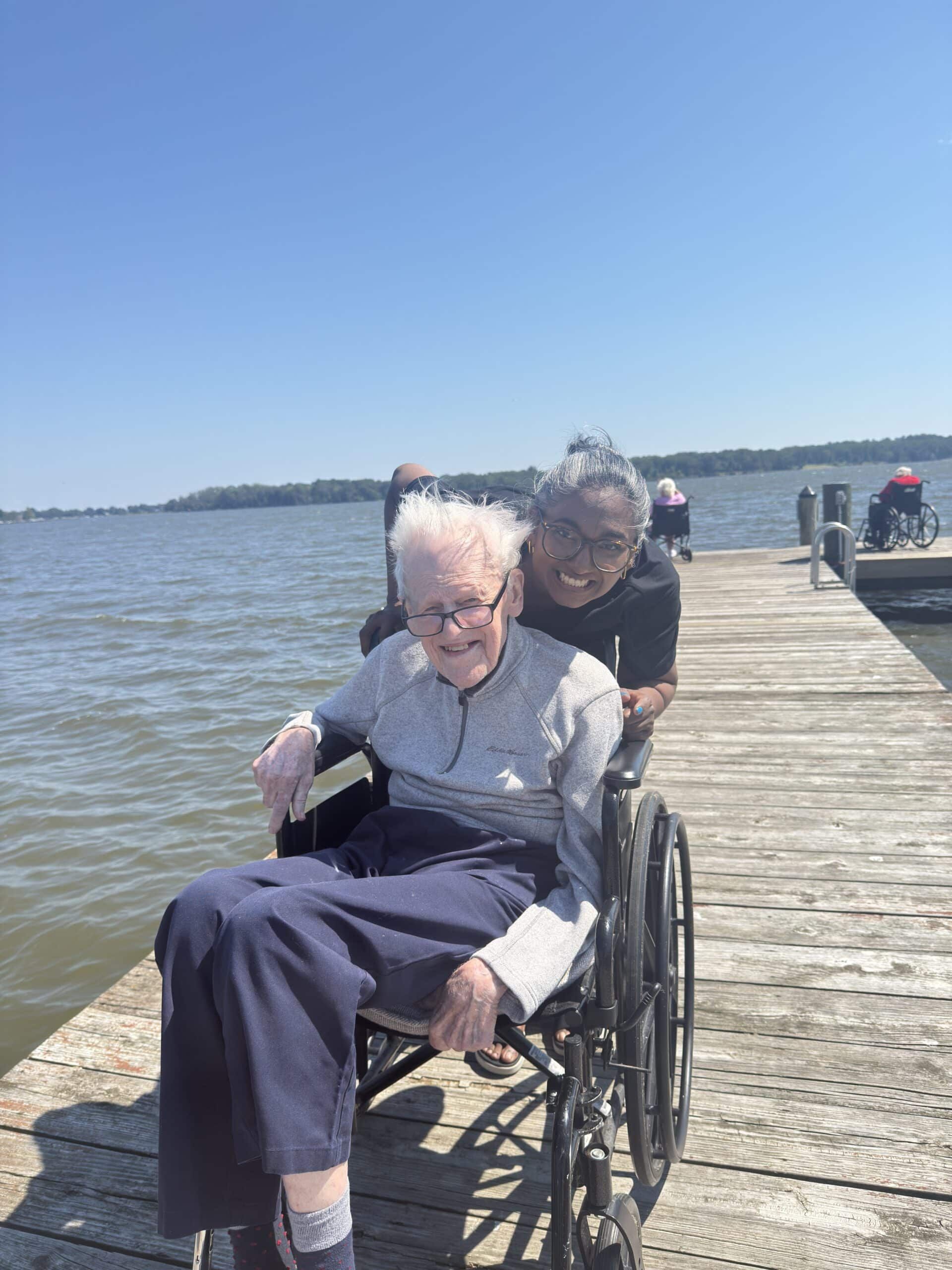 Woman in a wheelchair on a wooden pier by the water, with another person pushing from behind.
