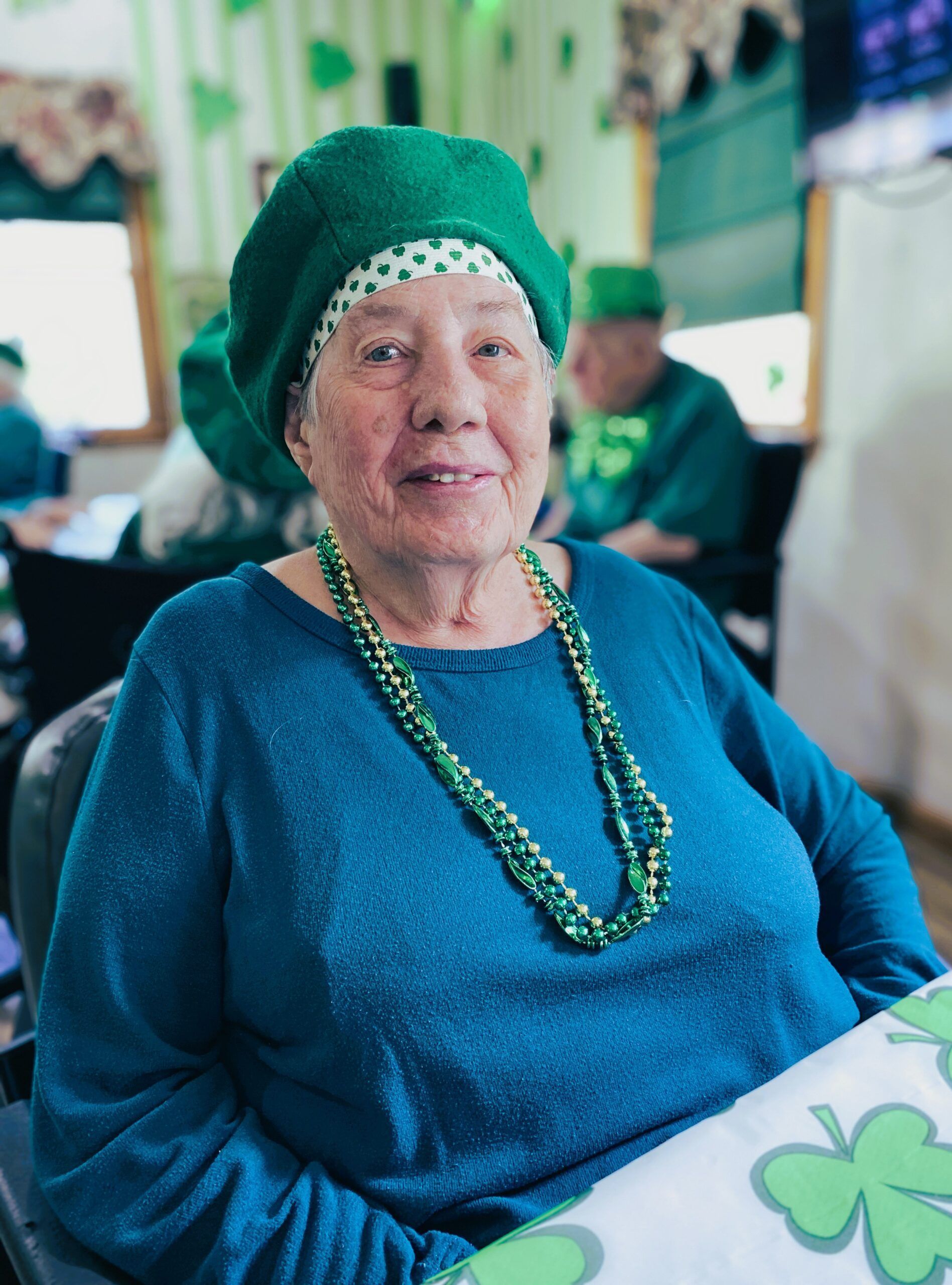 Elderly person in a green hat and blue sweater sits at a table with a beaded necklace, smiling gently.